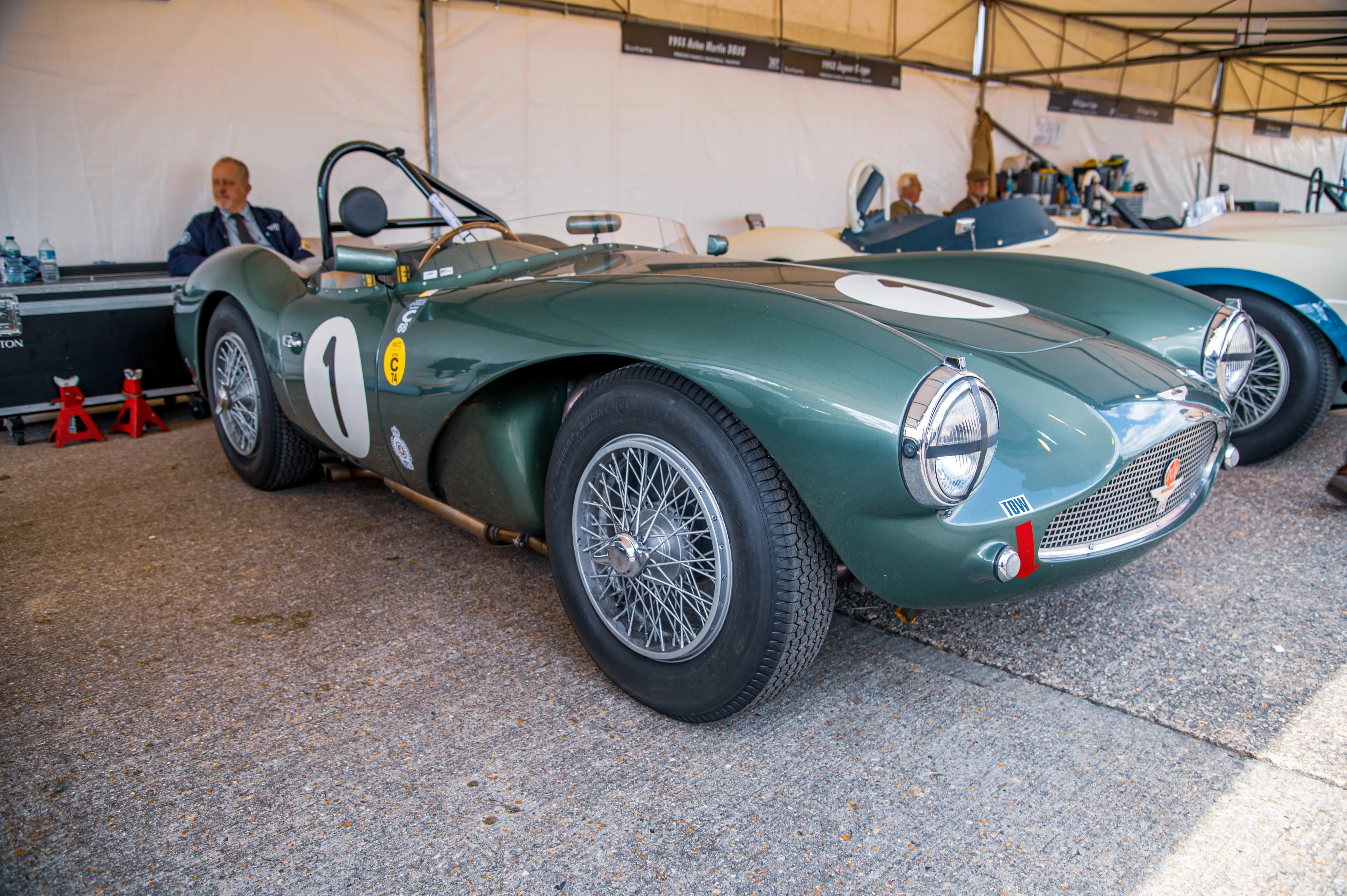 Close-up of a classic green Aston Martin DBR1 race car from the front, with a Jaguar C-Type visible behind it.