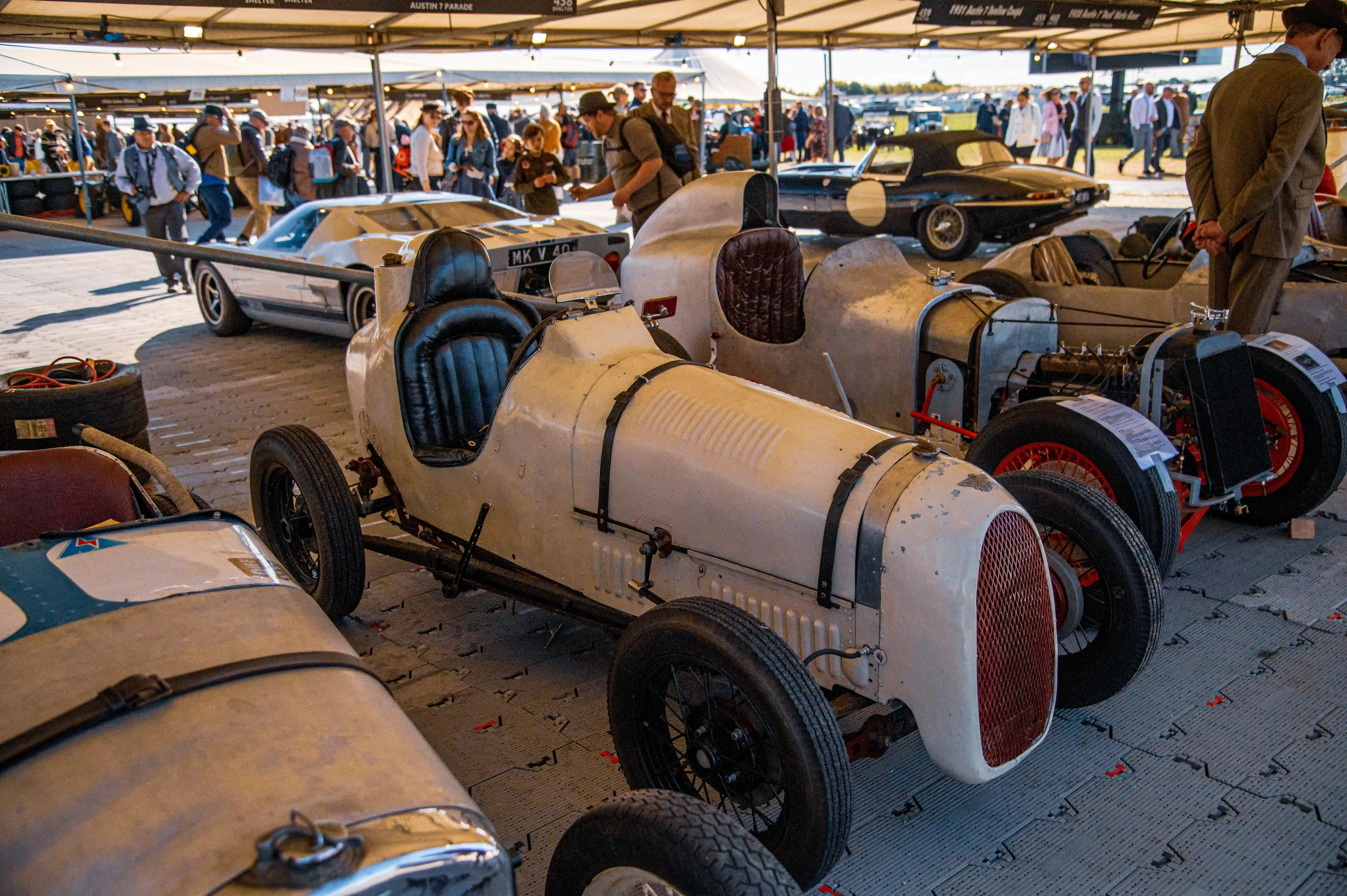 A selection of vintage and classic racing cars, some painted white and one with a black grille, are lined up outdoors under a covered structure. People are visible in the background, browsing the display.