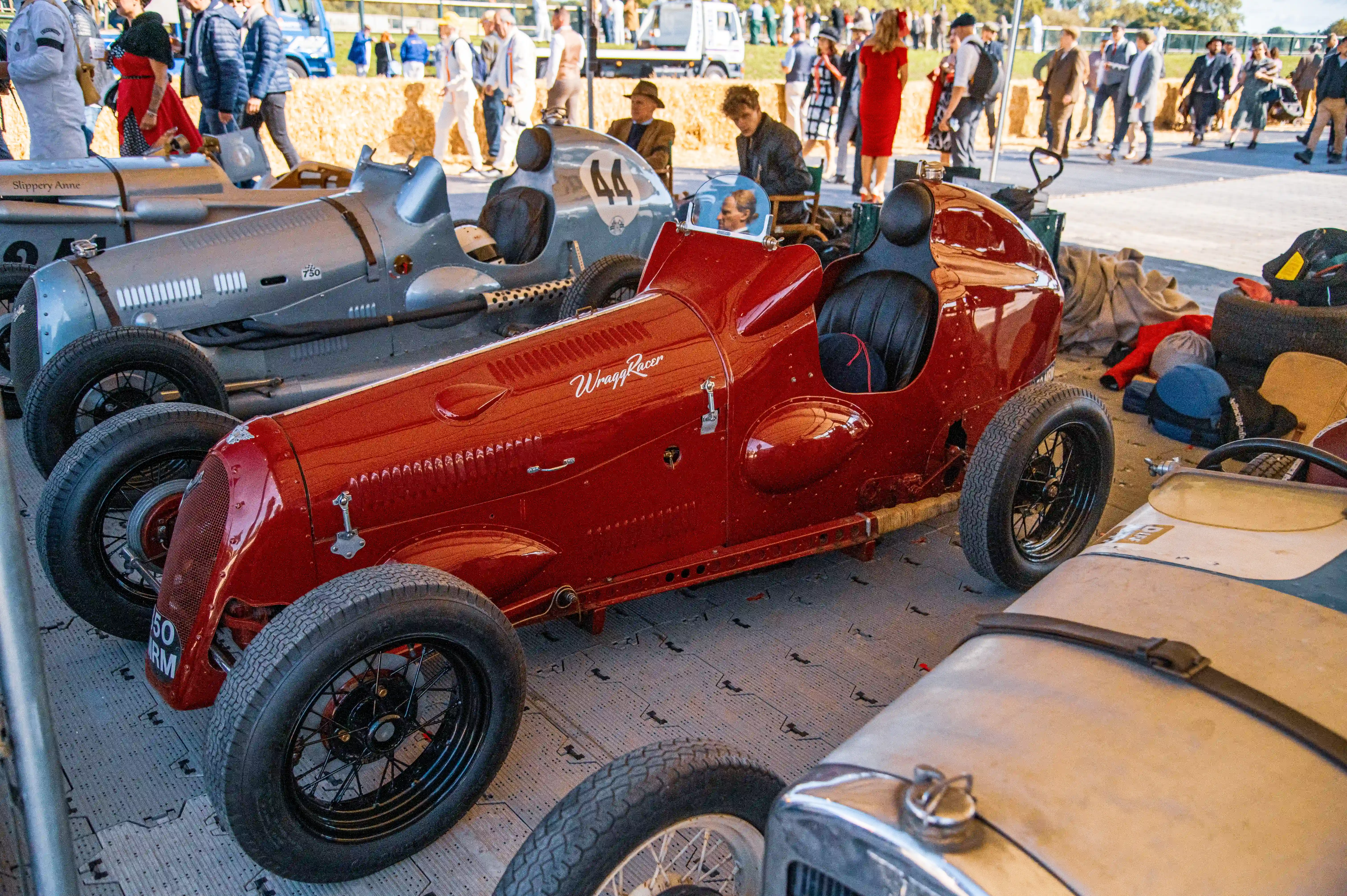 Close-up of a red vintage racing car with a silver racing car behind it at an outdoor event, with spectators in the background.