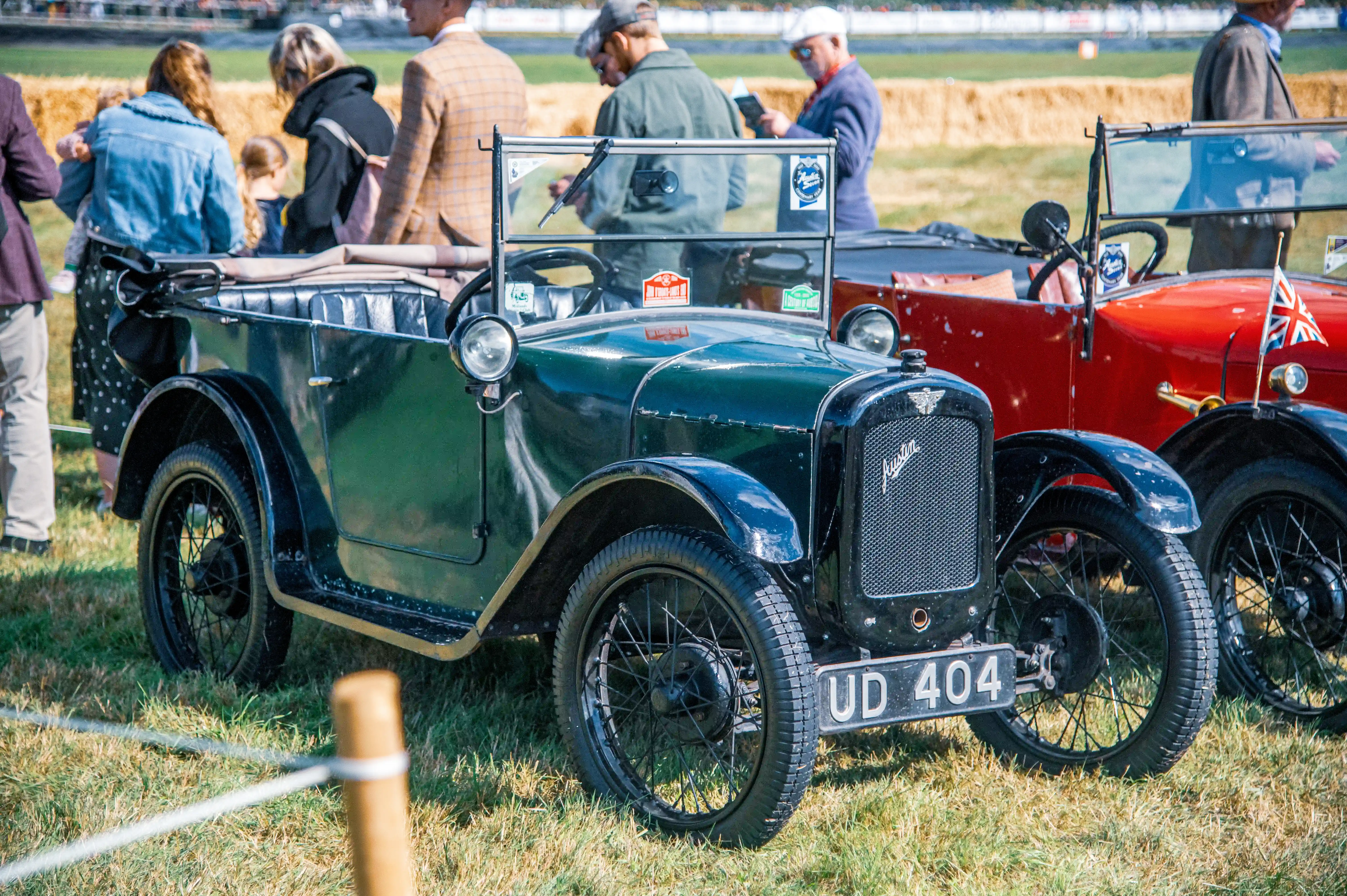 A dark green vintage car with a black soft-top roof, spoked wheels, and a chrome grille parked on a grassy field at a public event. The car’s license plate reads 'UD 404' and features a winged emblem on the front. In the background, a crowd of people, including adults and children, are gathered, some dressed in vintage attire. A red vintage car with a Union Jack flag is also visible