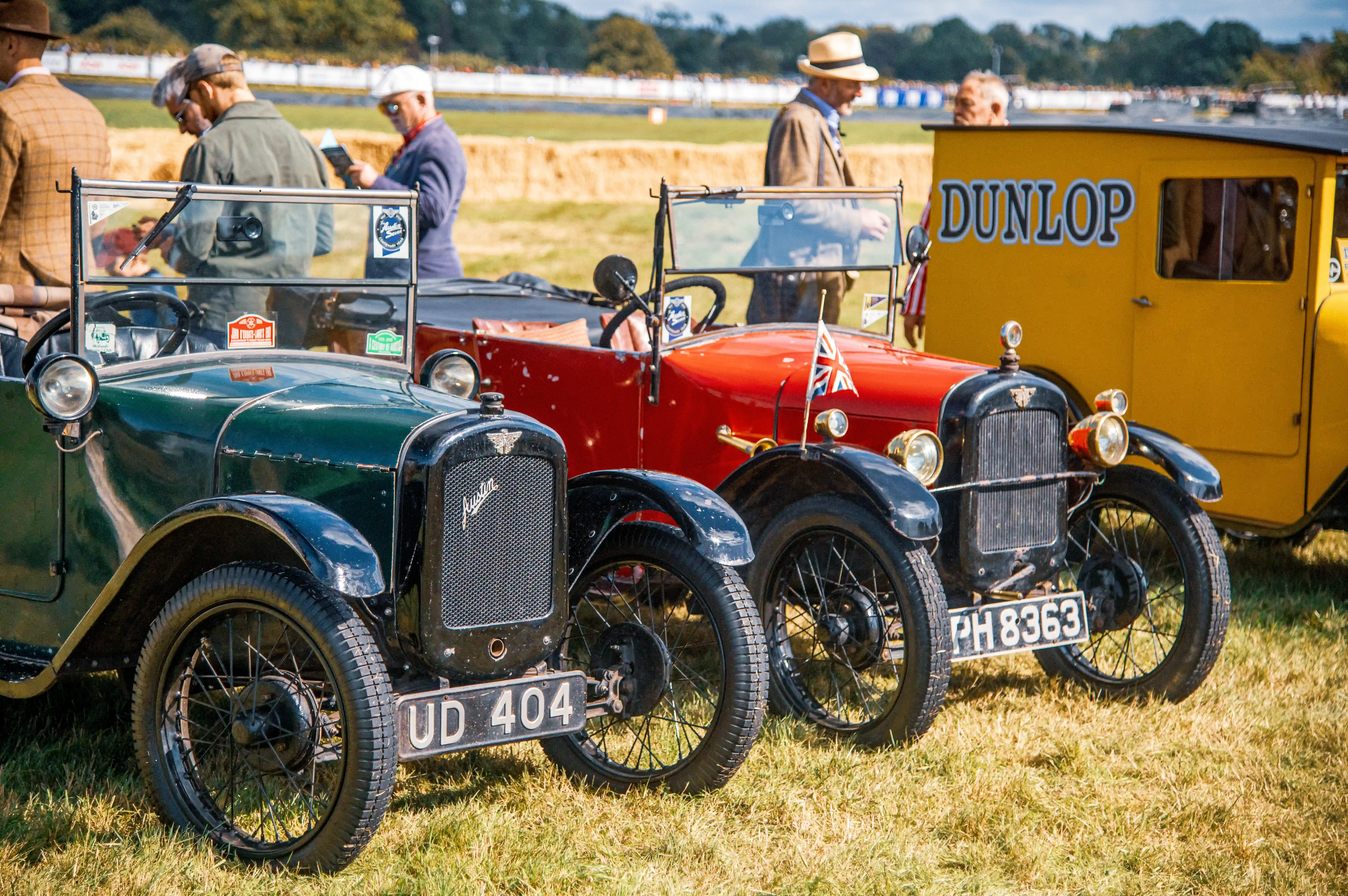 Close-up view of three vintage Austin cars parked side-by-side on a lawn, with people in the background. The cars are predominantly green and red, and a yellow van with 