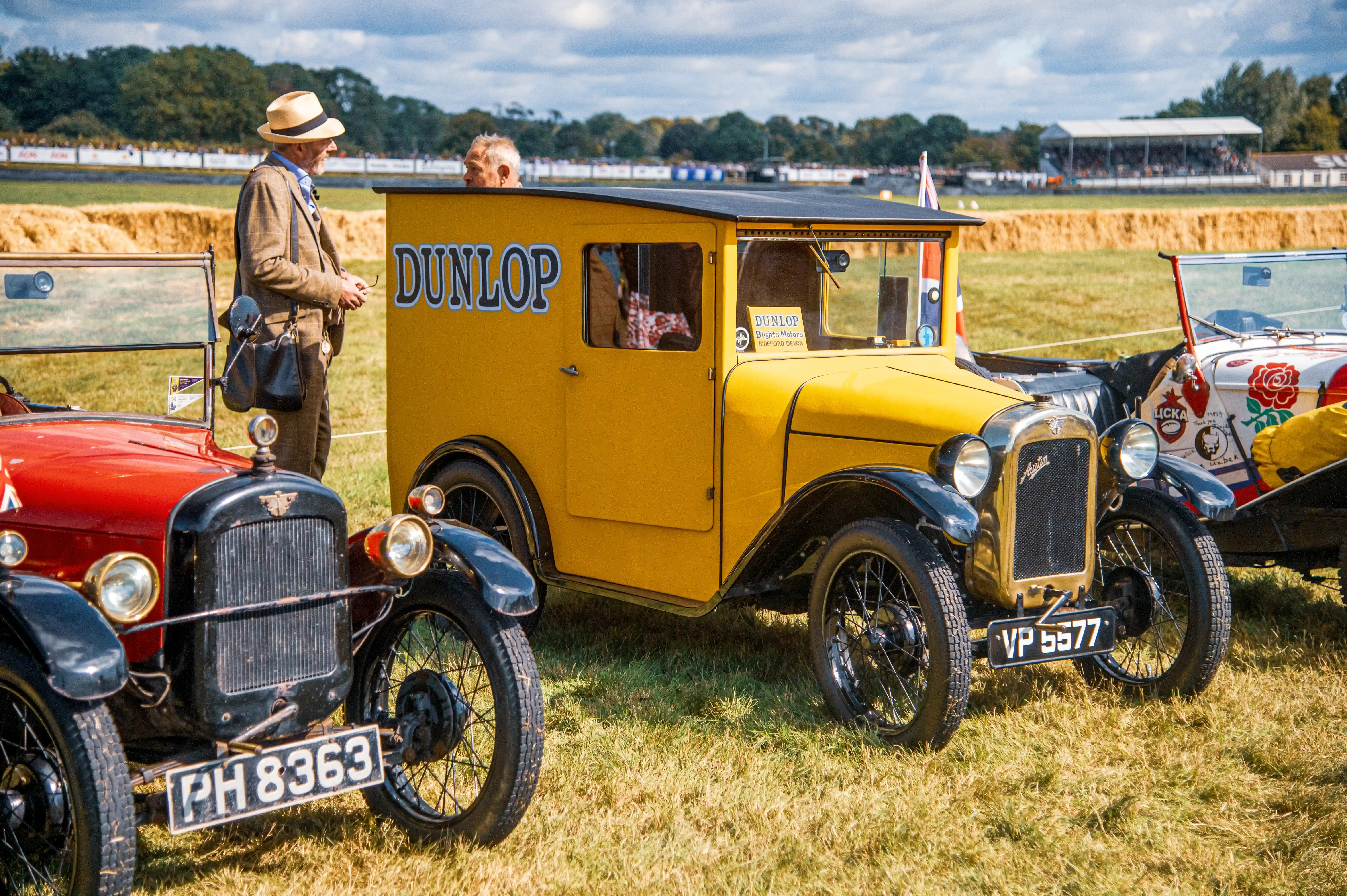 A bright yellow van with the Dunlop logo and a vintage red car are parked on a grassy field, with a spectator stand in the background under a cloudy sky.
