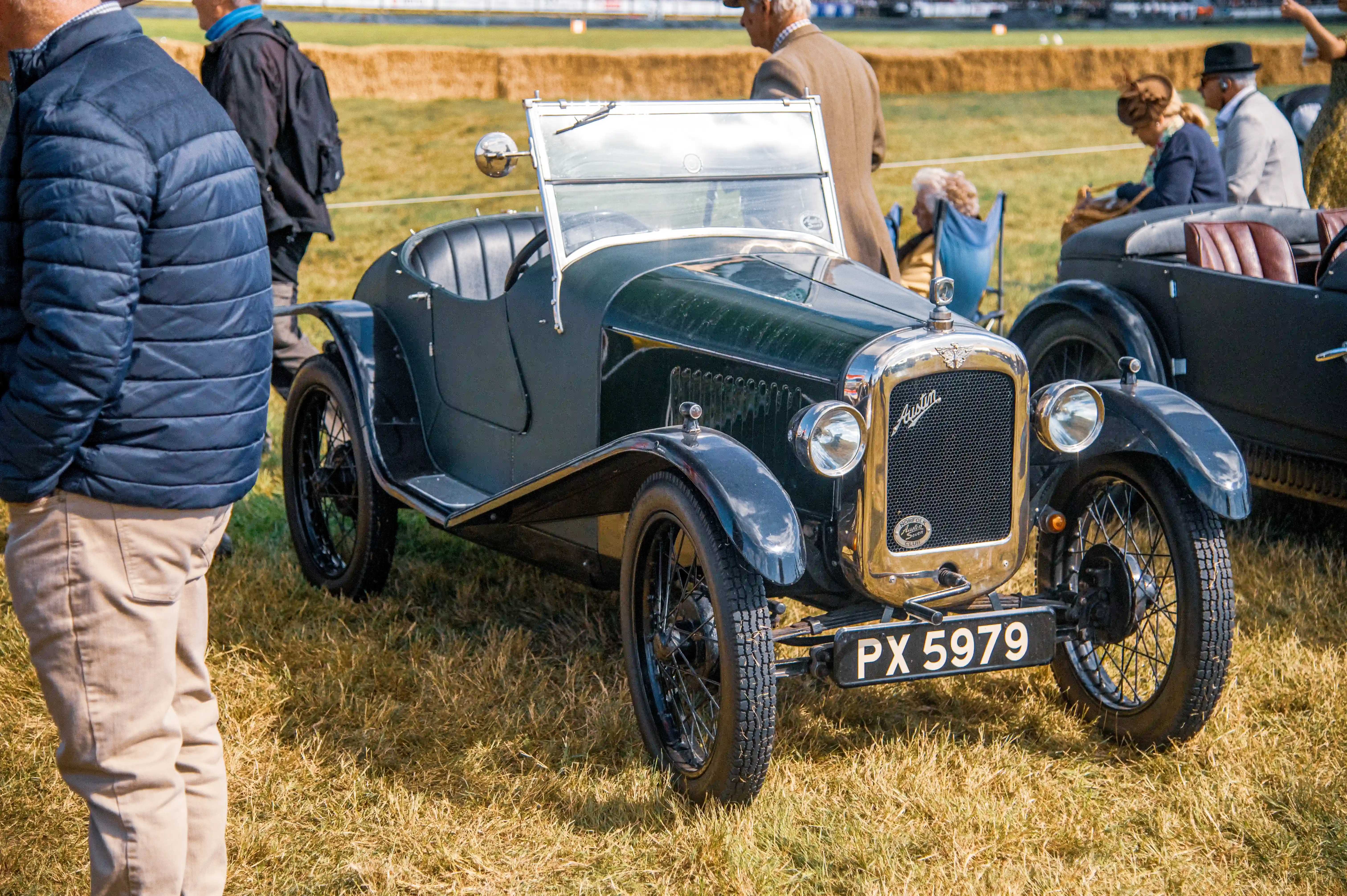 A dark green vintage Austin car with a chrome grille and wire wheels is parked on a grassy field. People are in the background.