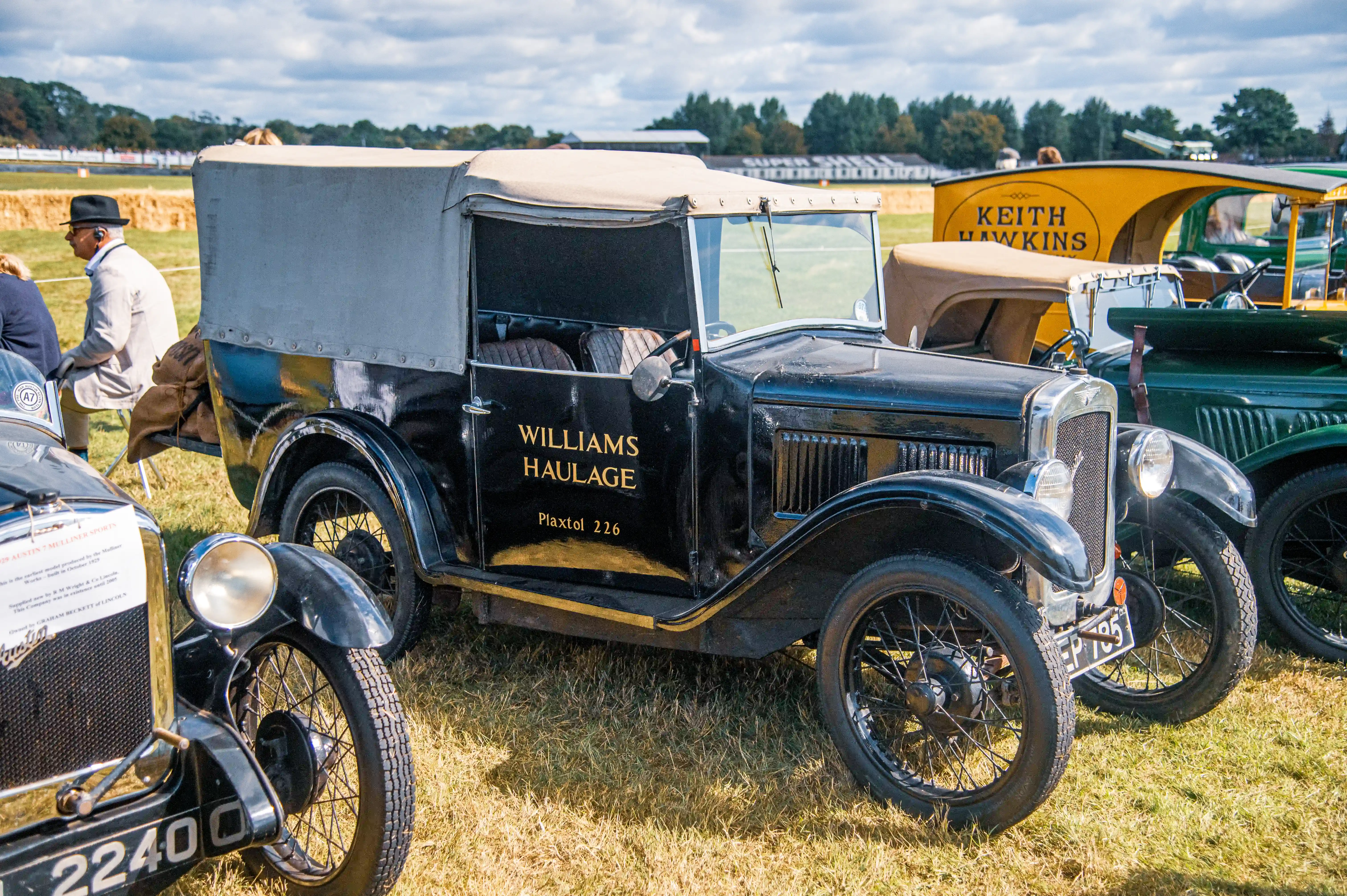 A black vintage van with a canvas roof is parked on grass next to other classic vehicles. The van has 'Williams Haulage' and 'Pixtel 226' written on its door. In the background, a yellow truck with 'Keith Hawkins' is visible.