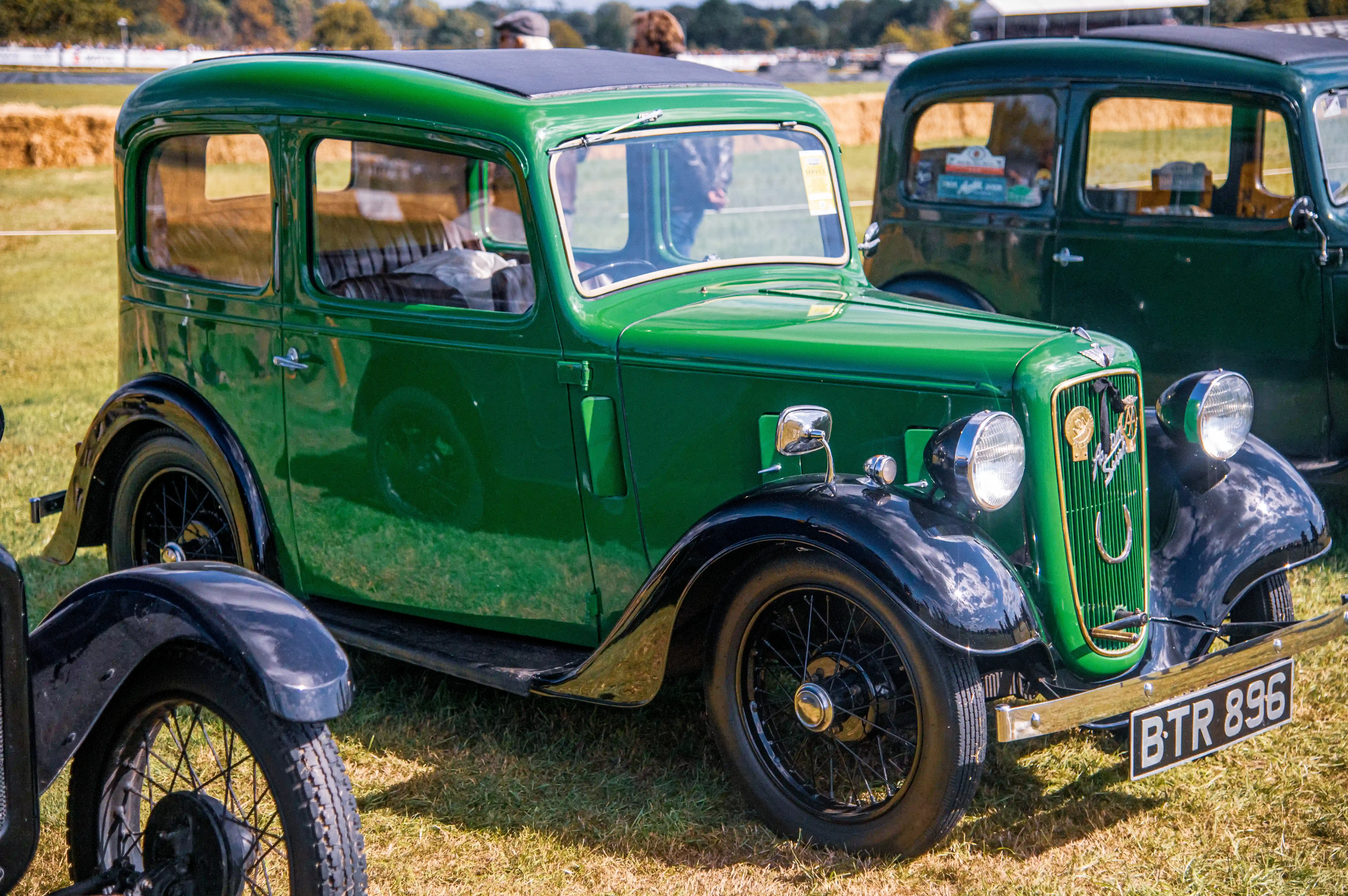A bright green vintage car with black fenders and running boards, parked on a grassy field at a classic car show. The car has a chrome grille, round headlamps, and a license plate reading 'BTR 896'. Other vintage cars and a crowd of people are visible in the background