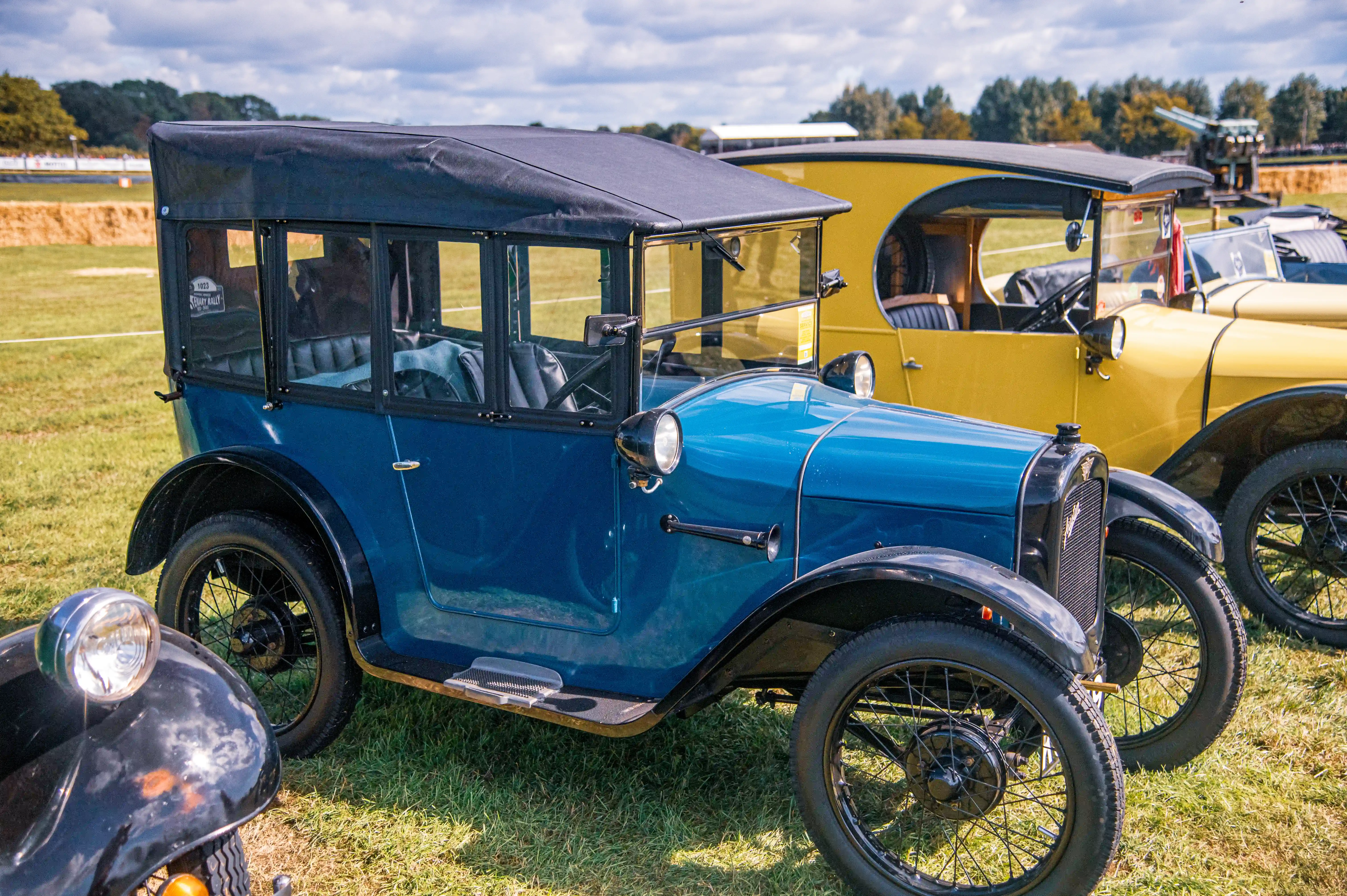 A close-up side view of a classic blue Austin Seven car with its hood up, parked on a lawn next to a yellow vintage car.
