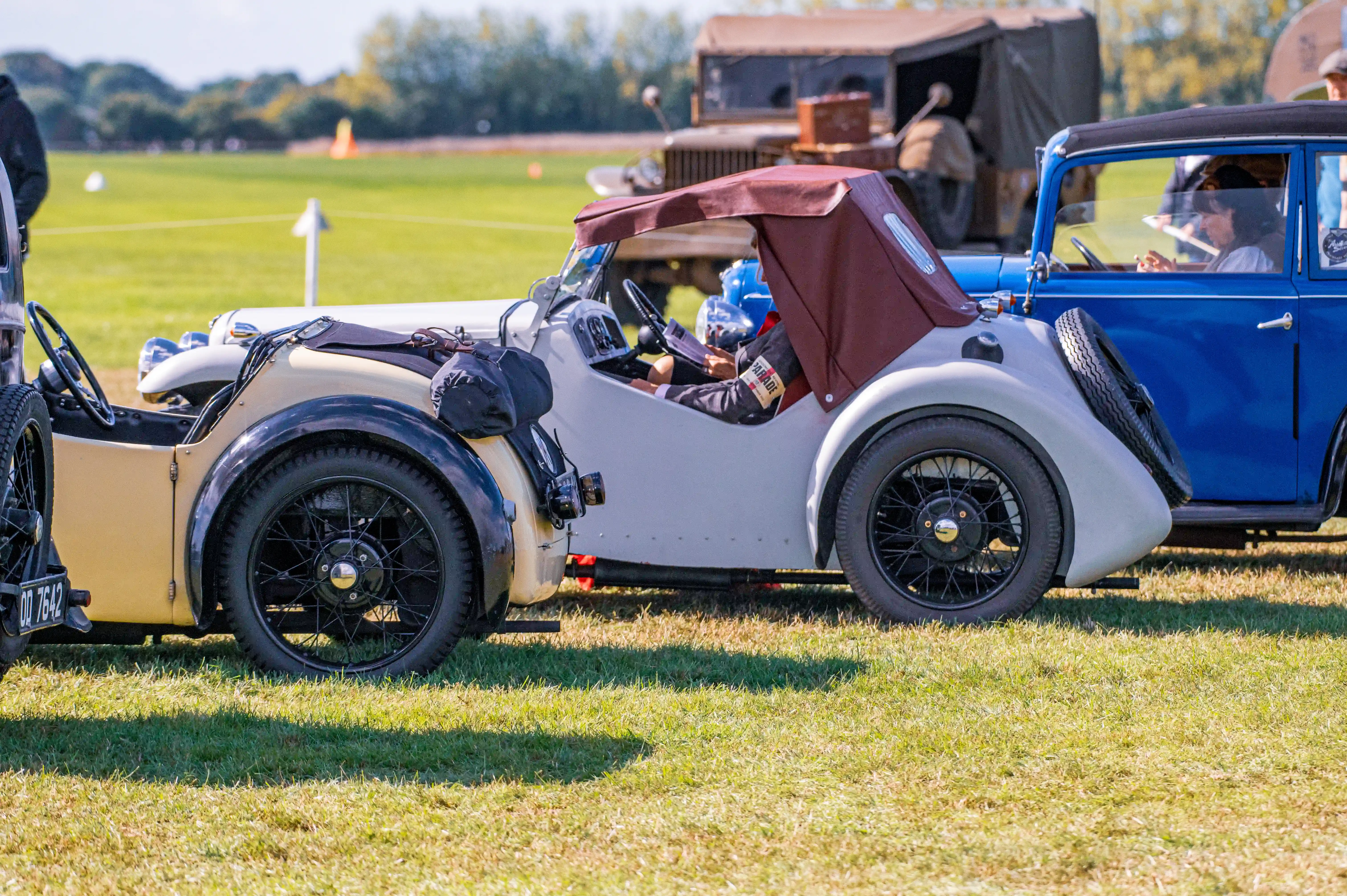 A side view of a vintage cream-coloured open-top car with a burgundy hood, parked on a green lawn next to a blue vintage car. Both cars have exposed wire wheels and appear to be part of a car show or event.