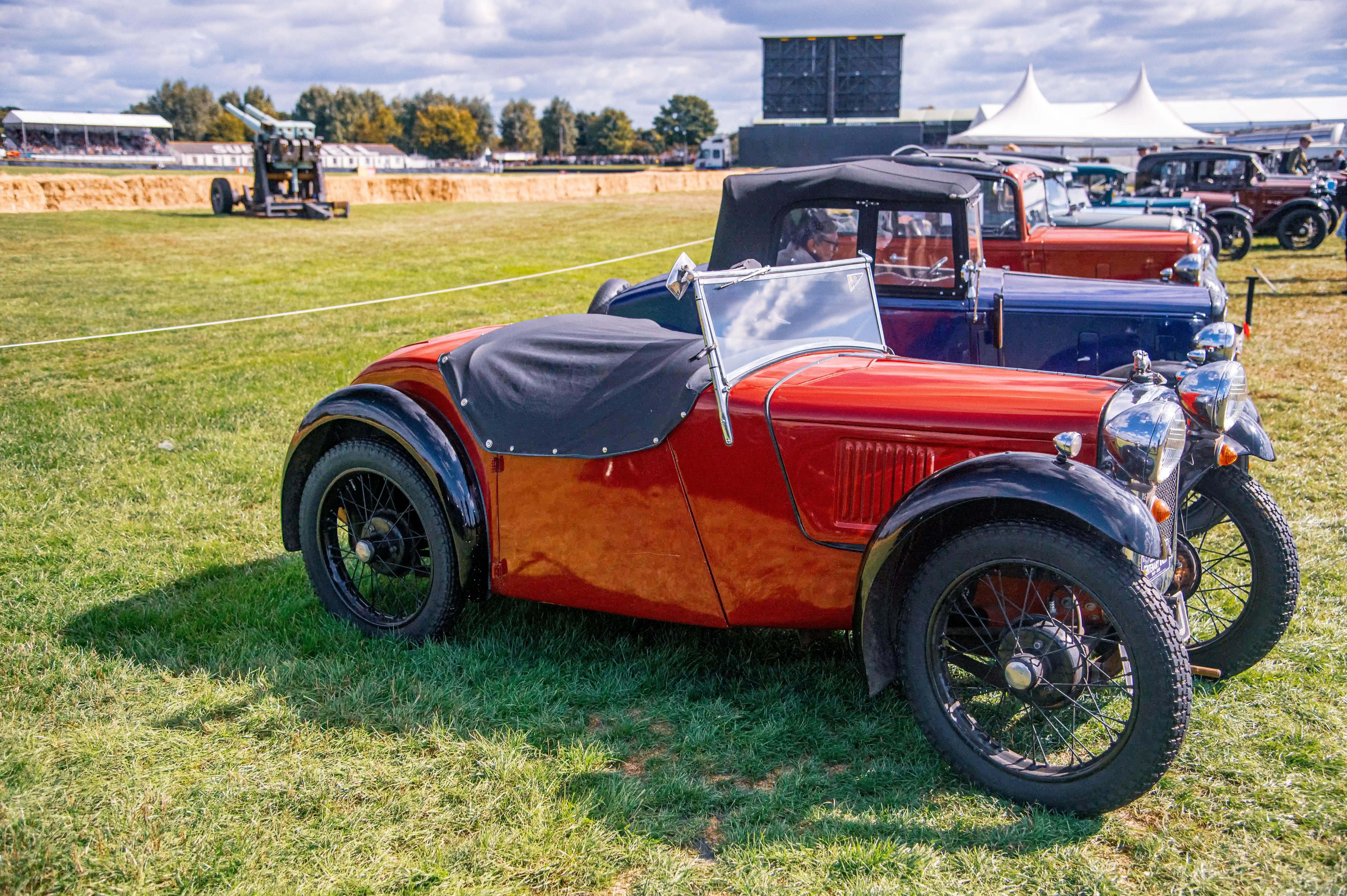 A close-up view of a red vintage convertible car with its black hood up, parked on a green lawn. More classic cars are lined up behind it.
