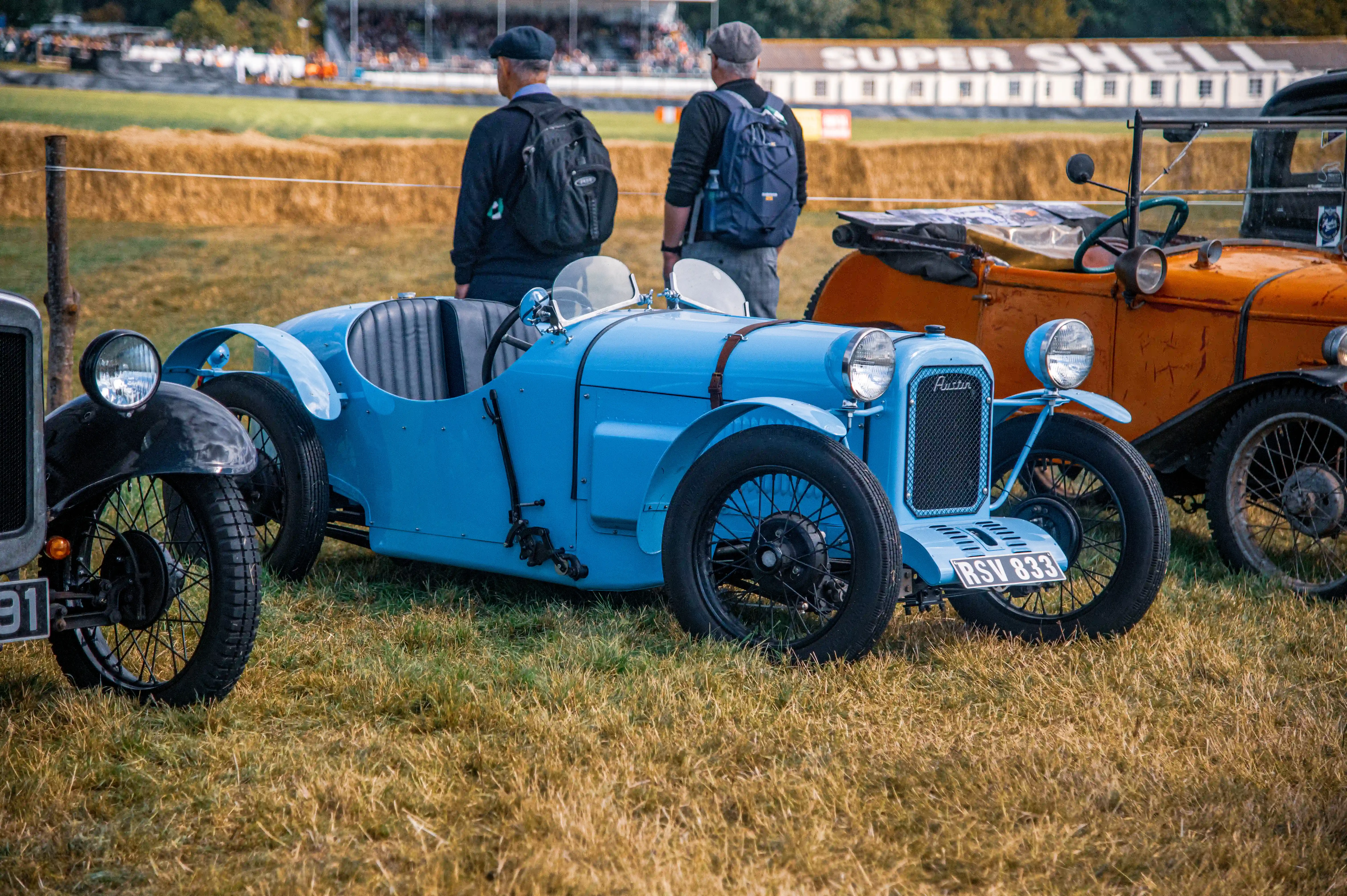 A light blue vintage Austin car with a streamlined body, chrome headlamps, and spoked wheels parked on a grassy field at a car show. The car’s license plate reads 'RSV 833' and features a black leather strap on the hood. In the background, an orange vintage car and two spectators with backpacks are visible, along with a 'Super Shell' sign and hay bales