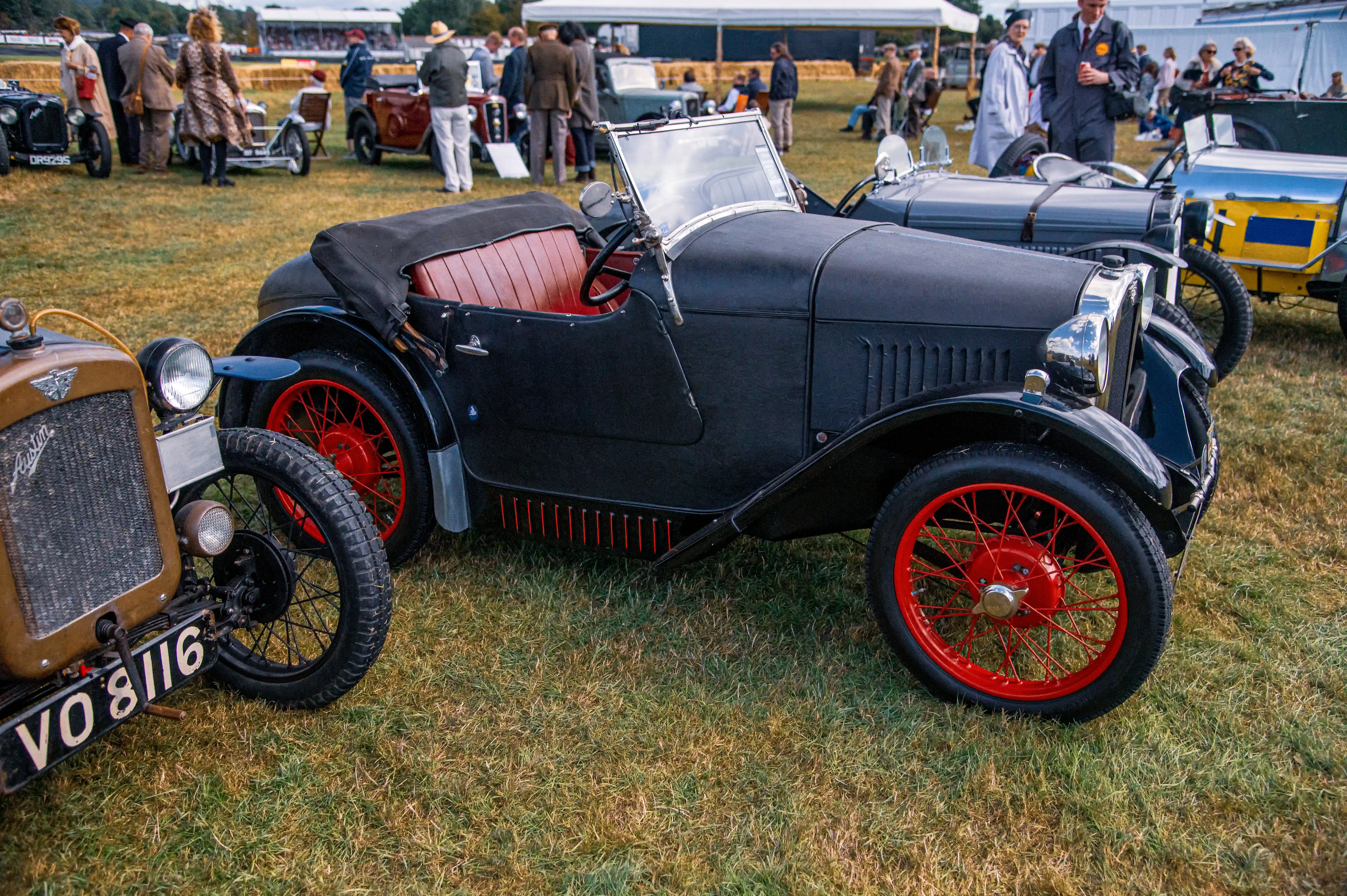 A black vintage car with red-spoked wheels and a tan soft-top roof parked on a grassy field at a public event. The car’s license plate reads 'VO 8116', and it features a chrome grille and round headlamps. In the background, a crowd of people is gathered, some dressed in vintage attire