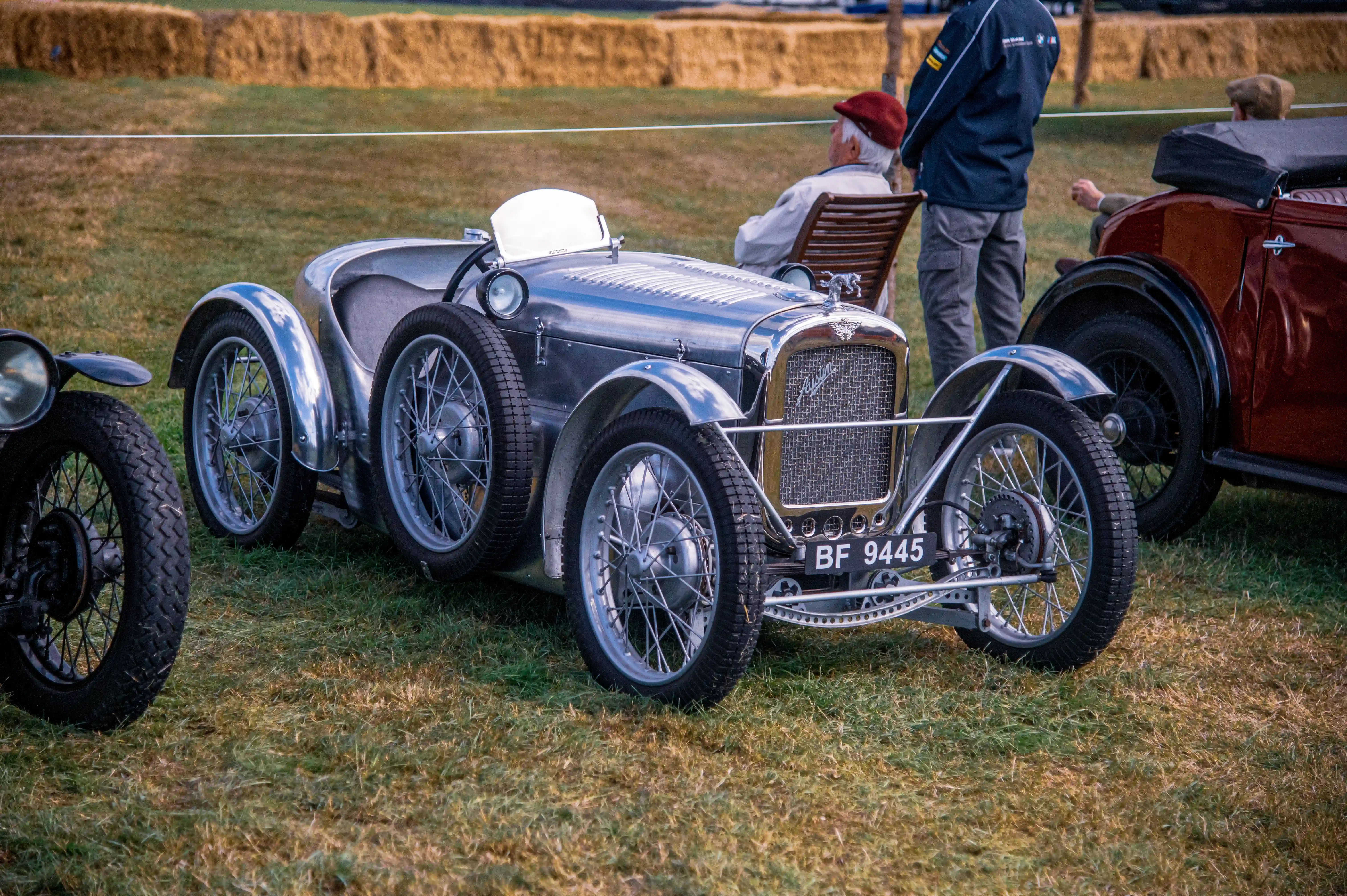 Close-up of a vintage silver Austin Seven racing car with polished metal bodywork and multiple spoked wheels, parked on grass. A red convertible car is visible to the right, and people in the background