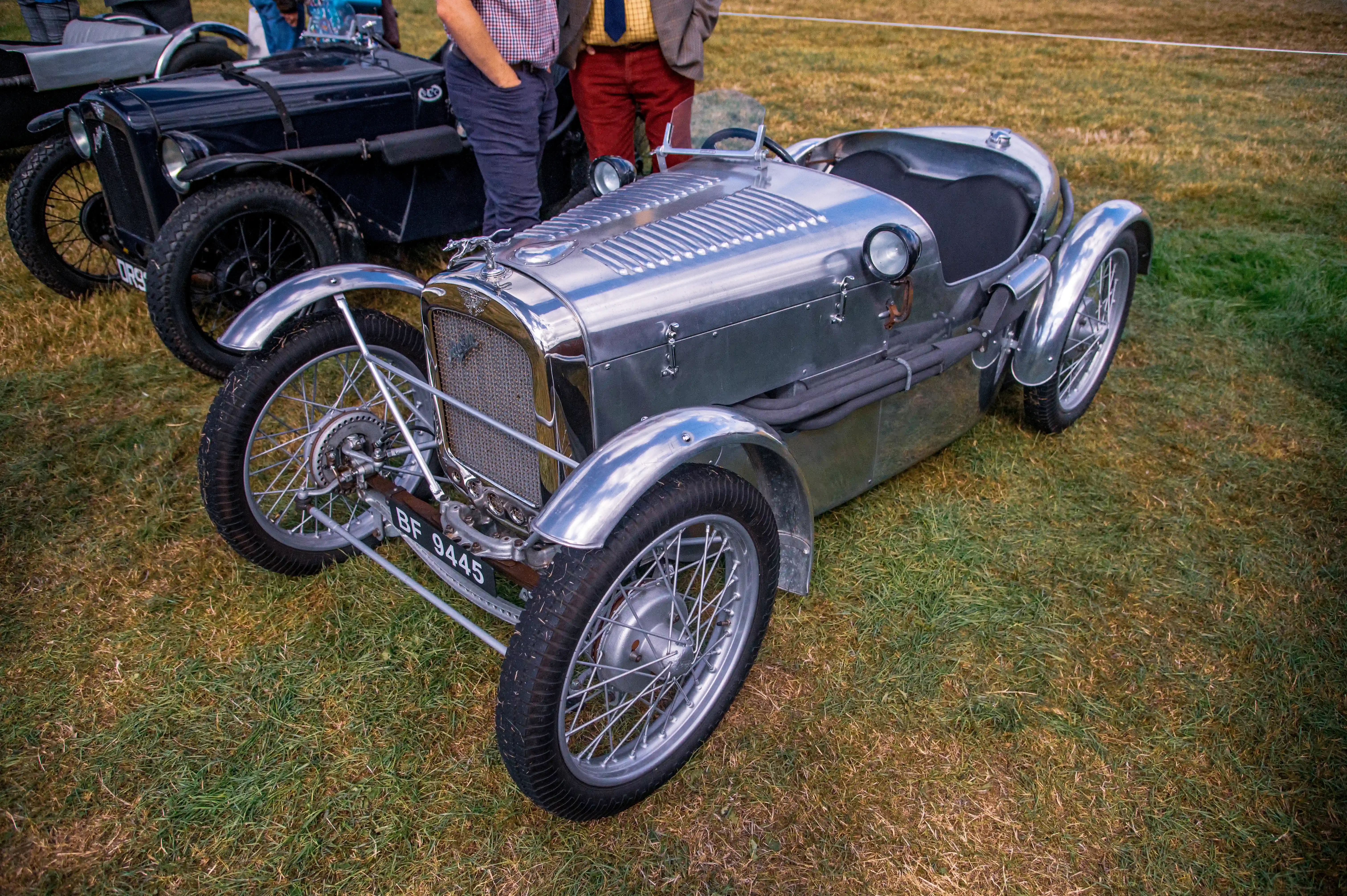 Close-up of a vintage silver Austin Seven racing car with polished metal bodywork and multiple spoked wheels, parked on grass. A red convertible car is visible to the right, and people in the background