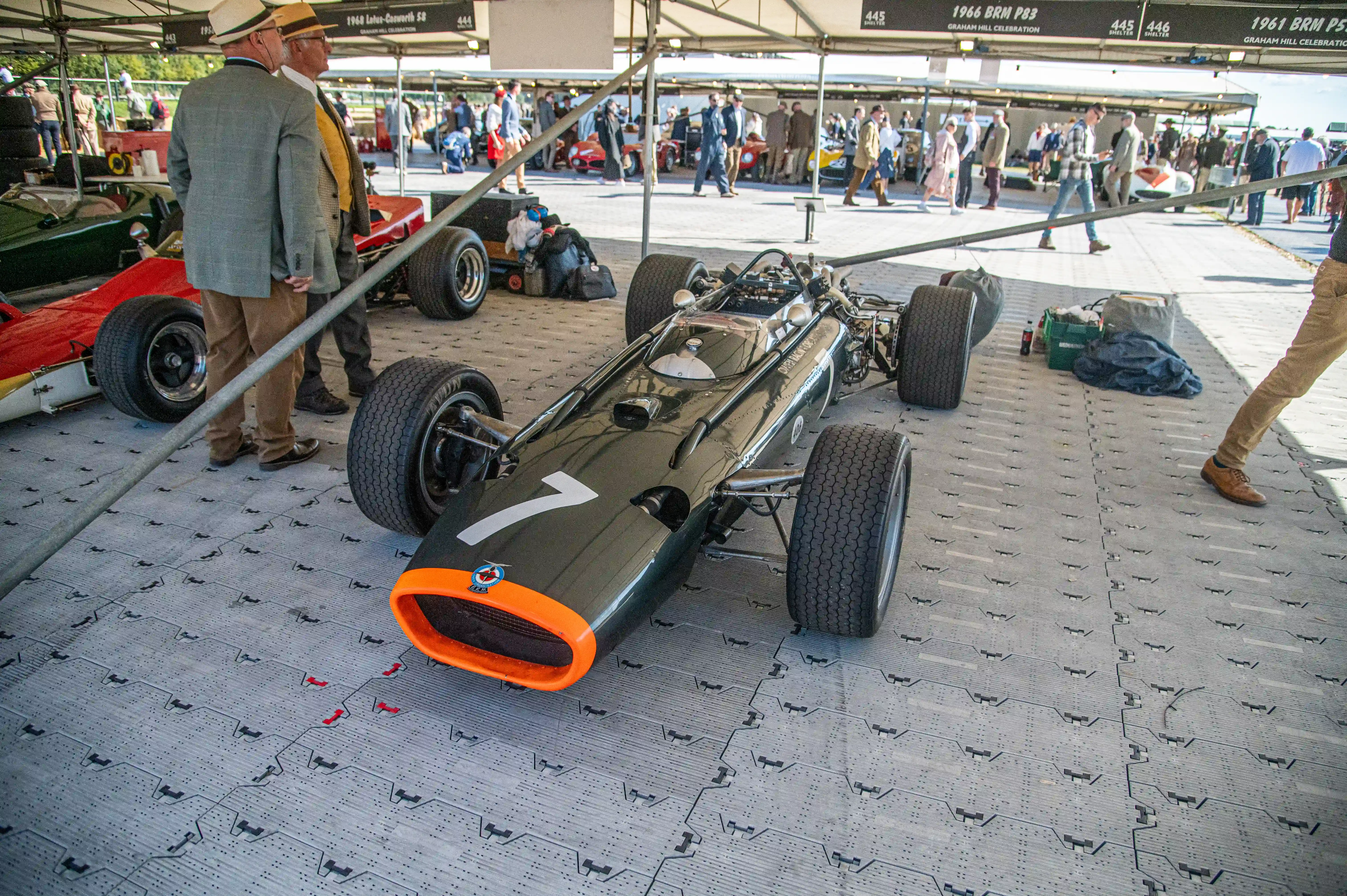 A vintage Formula 1 racing car, painted dark green with a bright orange front intake and the number 7 in white on its side, is displayed on a grey, patterned surface. Two men in hats and jackets stand behind it, while other cars and people are blurred in the background under a canopy.