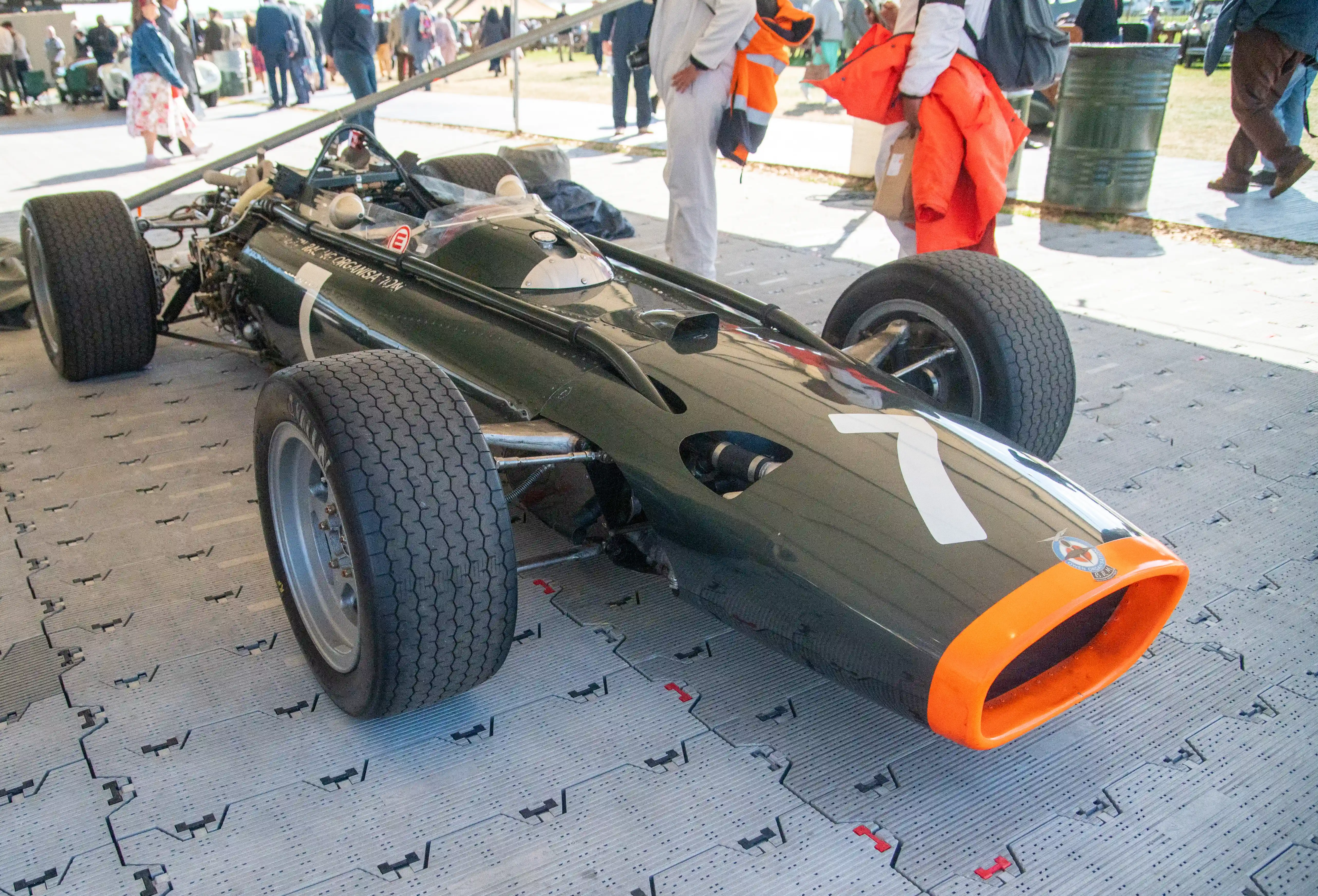 A vintage Formula 1 racing car, painted dark green with a bright orange front intake and the number 7 in white on its side, is displayed on a grey, patterned surface. Two men in hats and jackets stand behind it, while other cars and people are blurred in the background under a canopy.