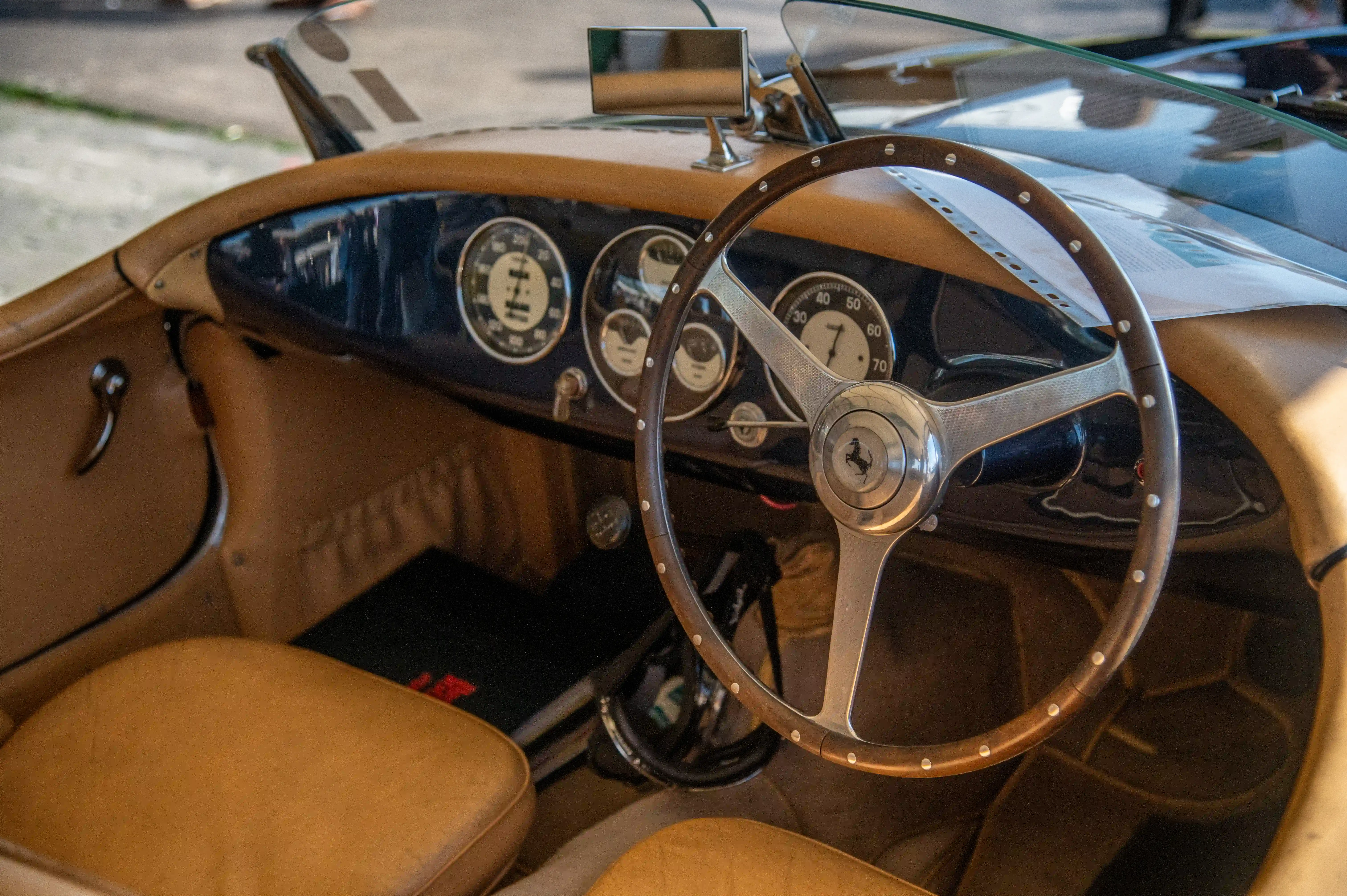 The interior of a classic Ferrari sports car, featuring a wooden steering wheel with a central Ferrari prancing horse emblem, multiple analogue gauges, and tan leather seats.