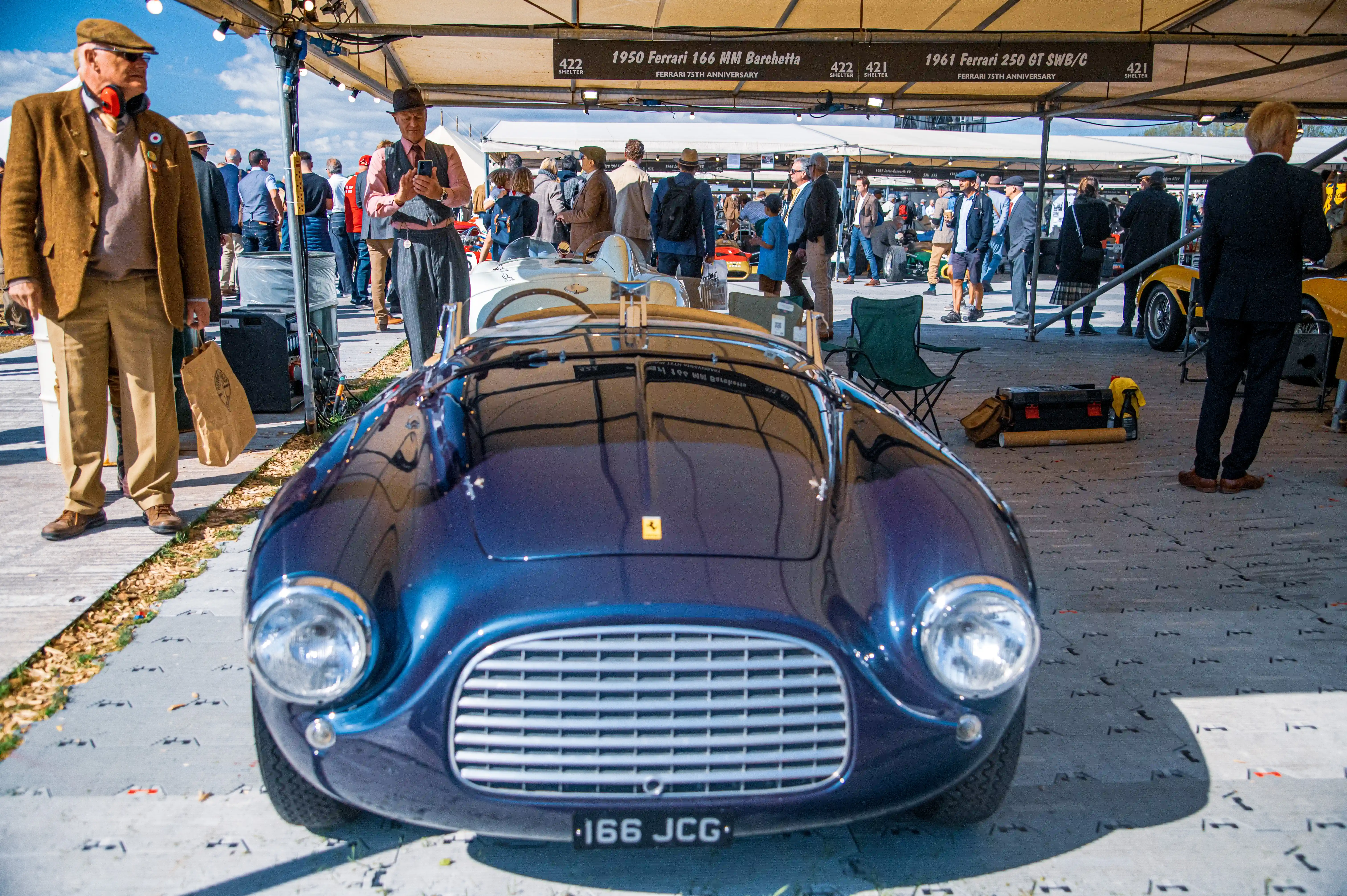 Front view of a gleaming dark blue vintage Ferrari with a prominent silver grille and round headlights, parked outdoors under a tented area. Several people in the background are looking at classic cars.