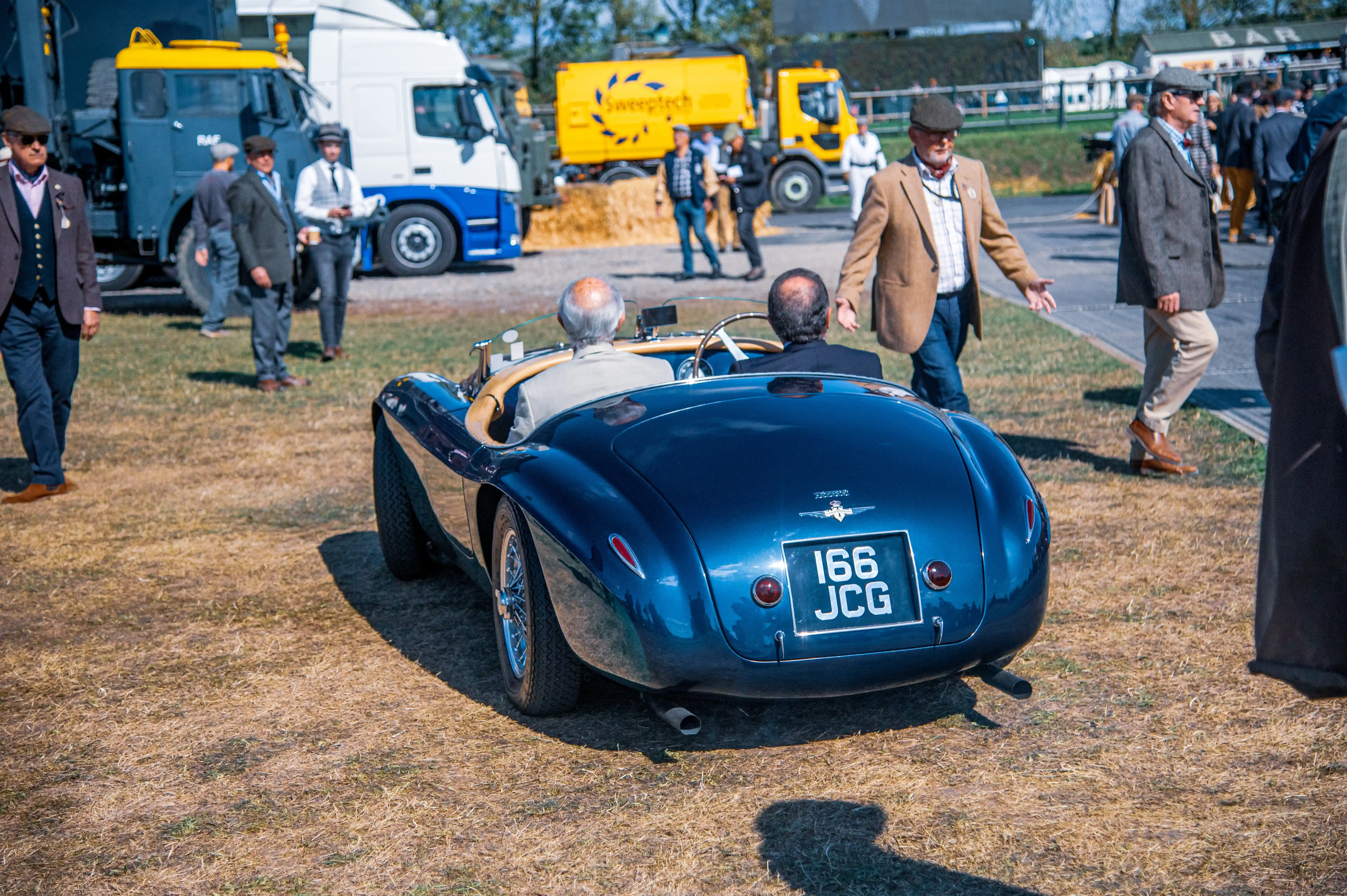 A dark blue vintage convertible sports car is viewed from the rear, with two men sitting inside. Several people in smart casual clothing are standing around the car and in the background, where trucks are parked.
