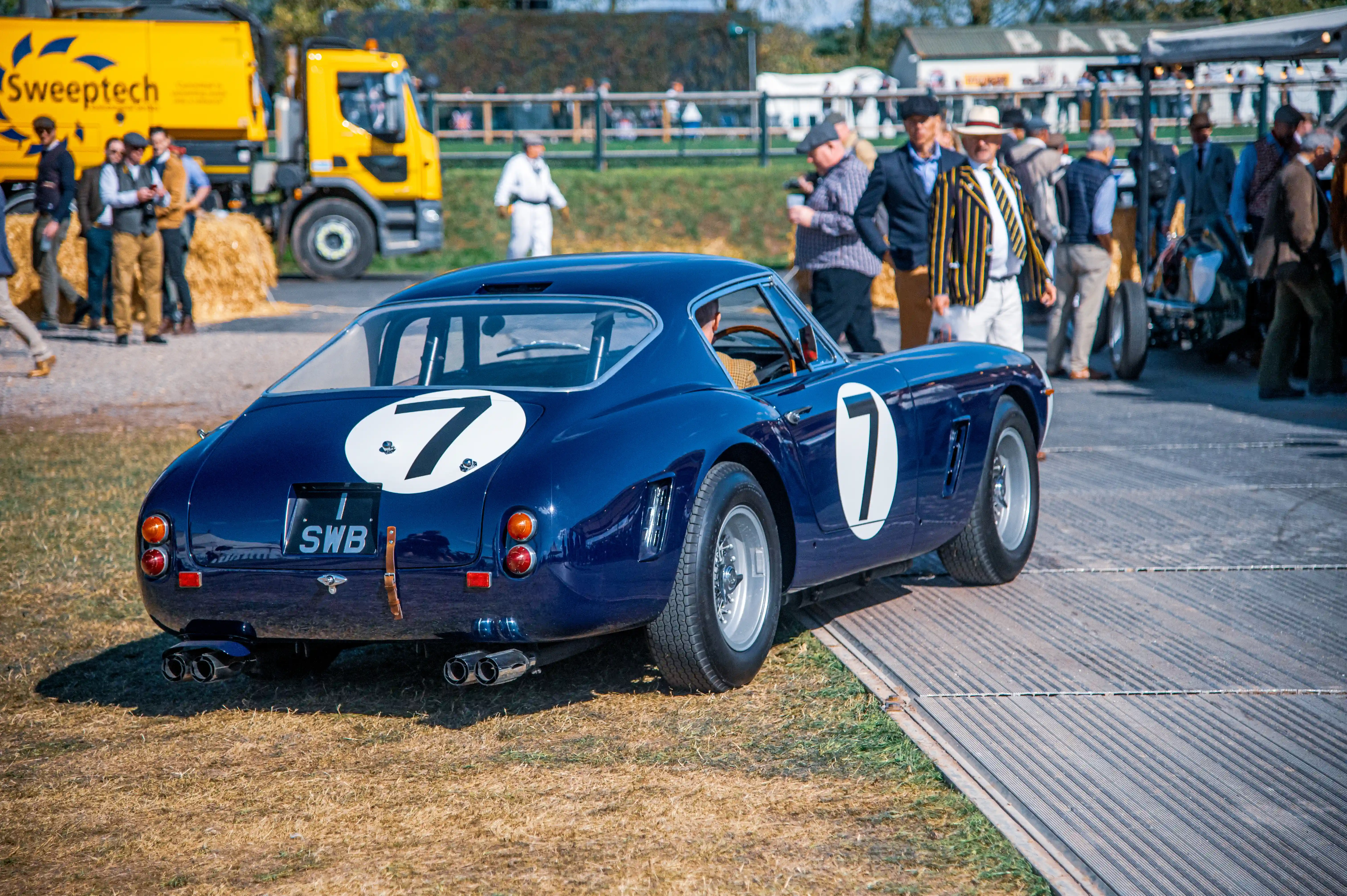 Rear three-quarter view of a dark blue classic Ferrari 250 GT Berlinetta SWB with white racing number '7' decals on the boot and side. The car has chrome quad exhaust pipes and a UK license plate reading 'SWB'. In the background, a yellow Sweeptech lorry and people are visible.