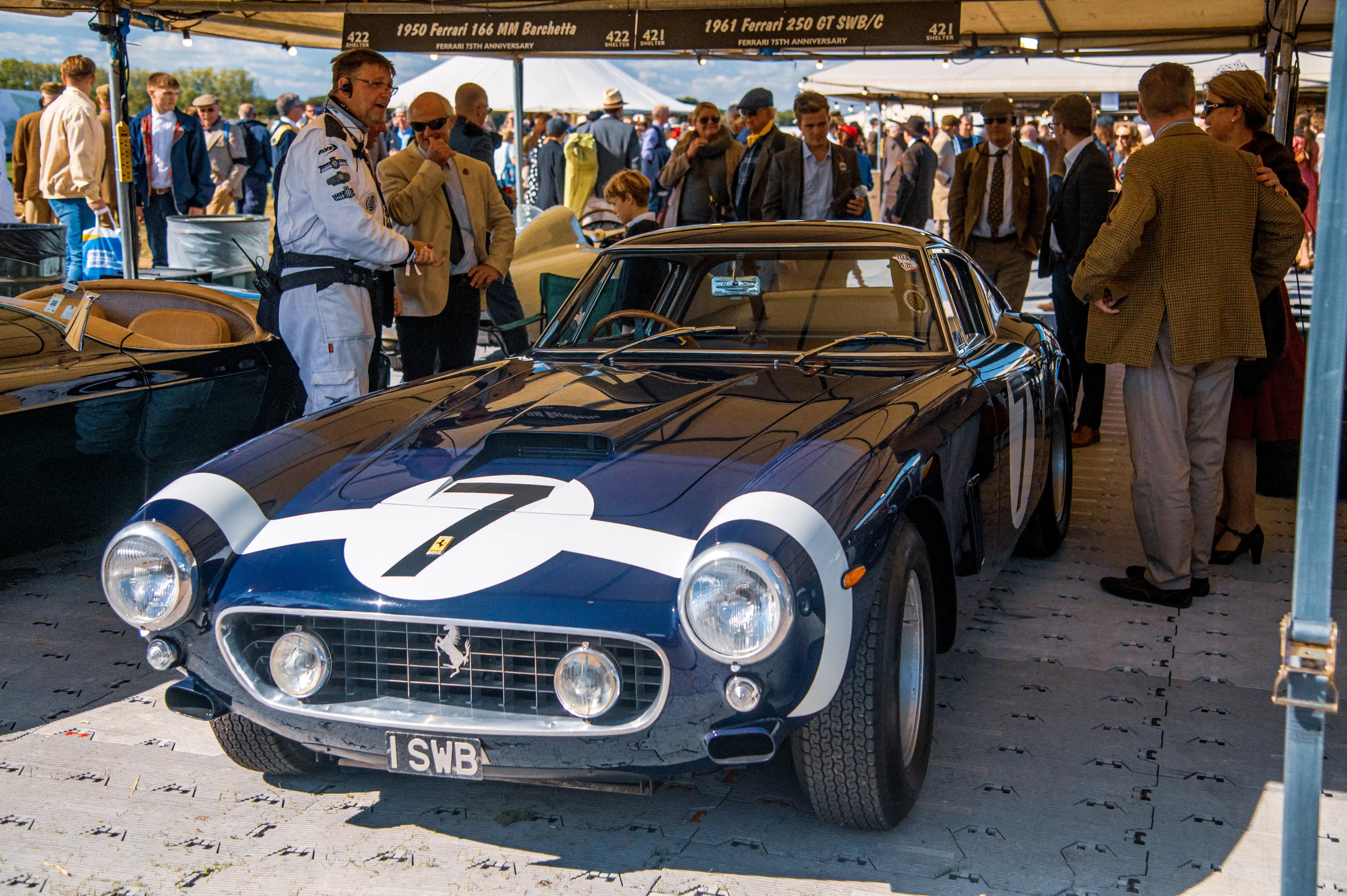A close-up front view of a vintage dark blue Ferrari 250 GT SWB/C with white racing stripes and the number 7 visible, parked on a grey tiled floor under a tent. People in smart attire are visible in the background.