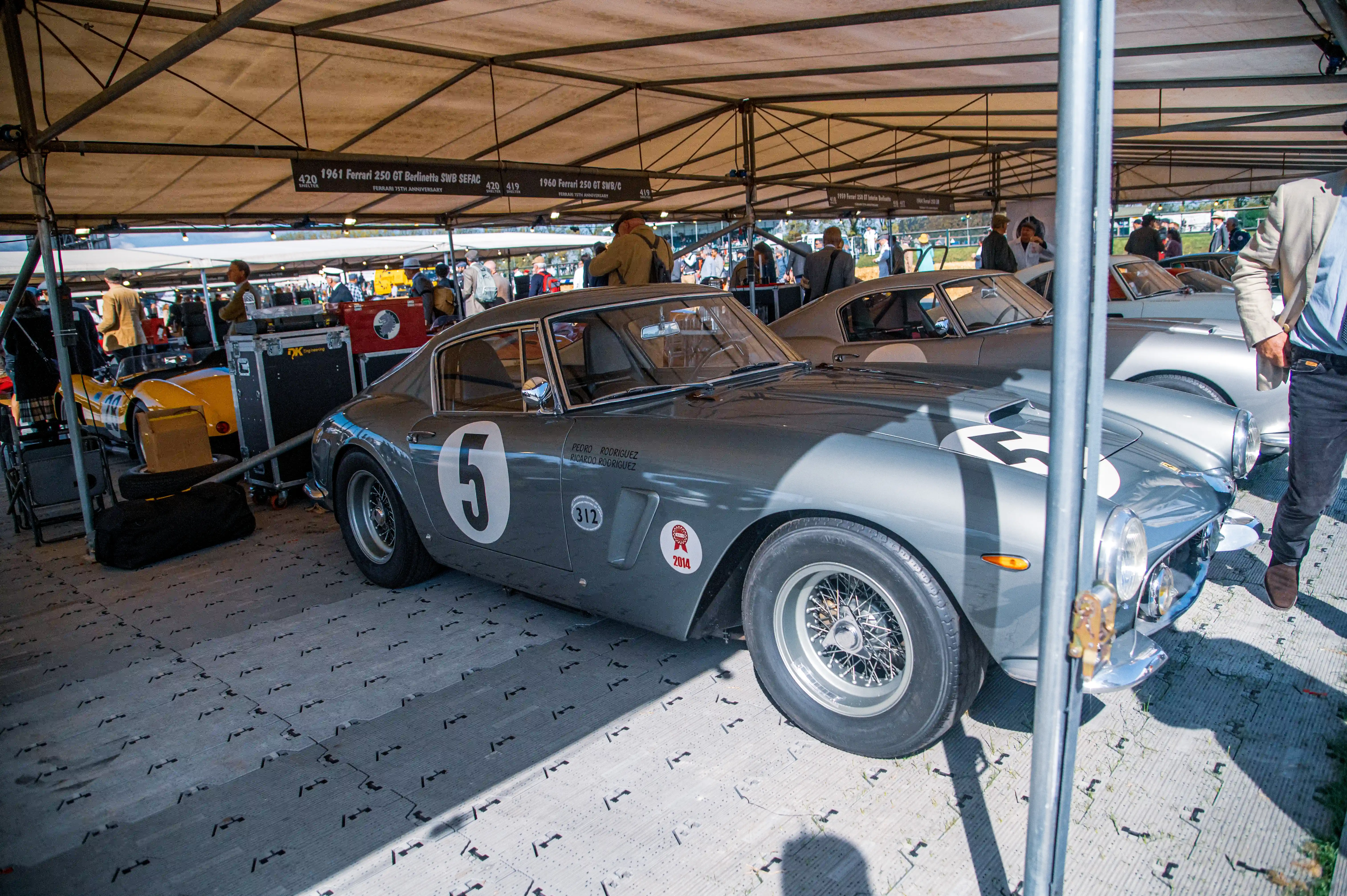 A grey 1961 Ferrari 250 GT Berlinetta SWB SEFAC with the number 5 on the door is parked under a tent. Other vintage cars and people are visible in the background.