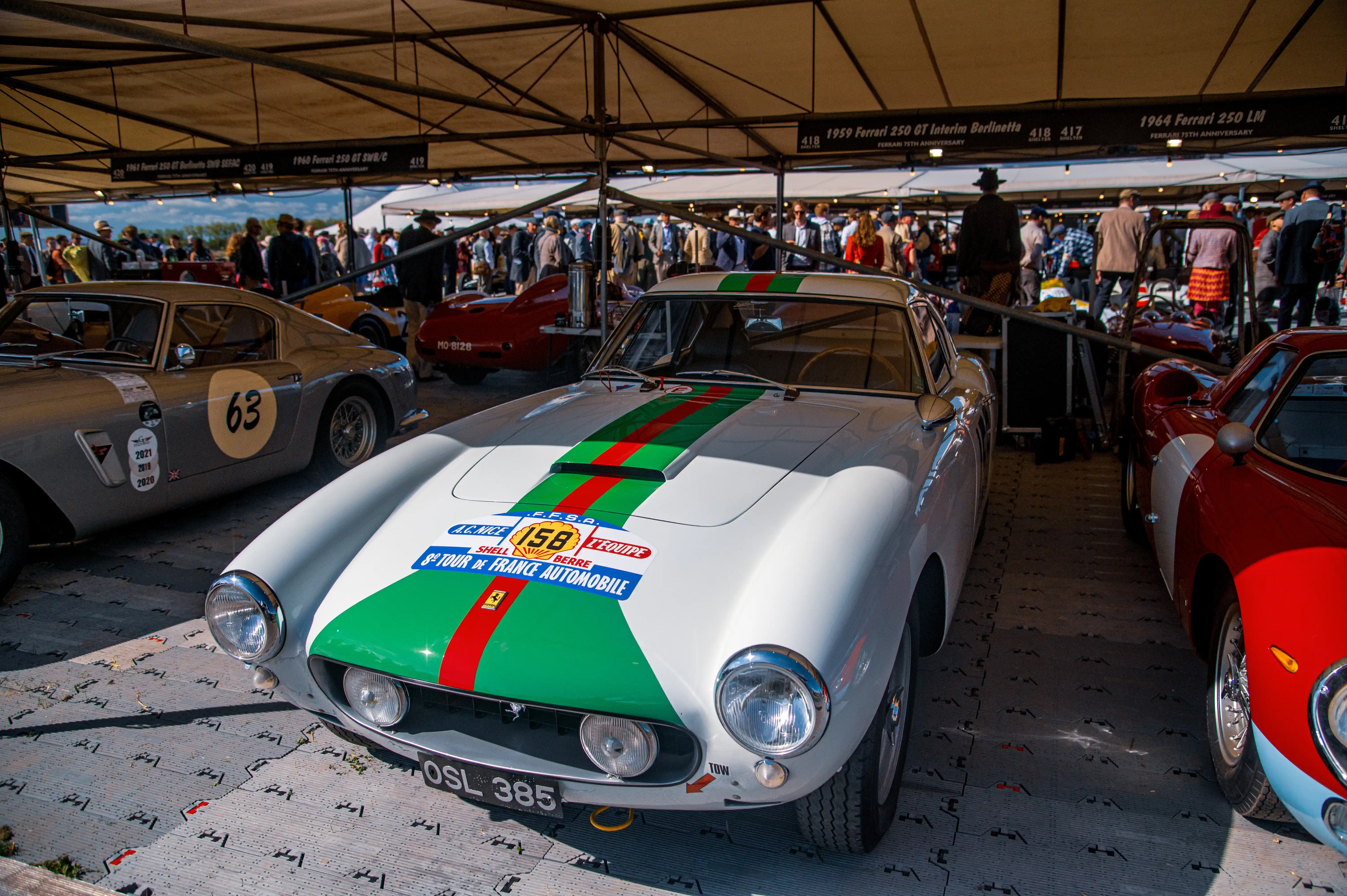 A white classic Ferrari 250 GT with distinctive green and red racing stripes is parked in the foreground. It has decals on its bonnet indicating participation in the 