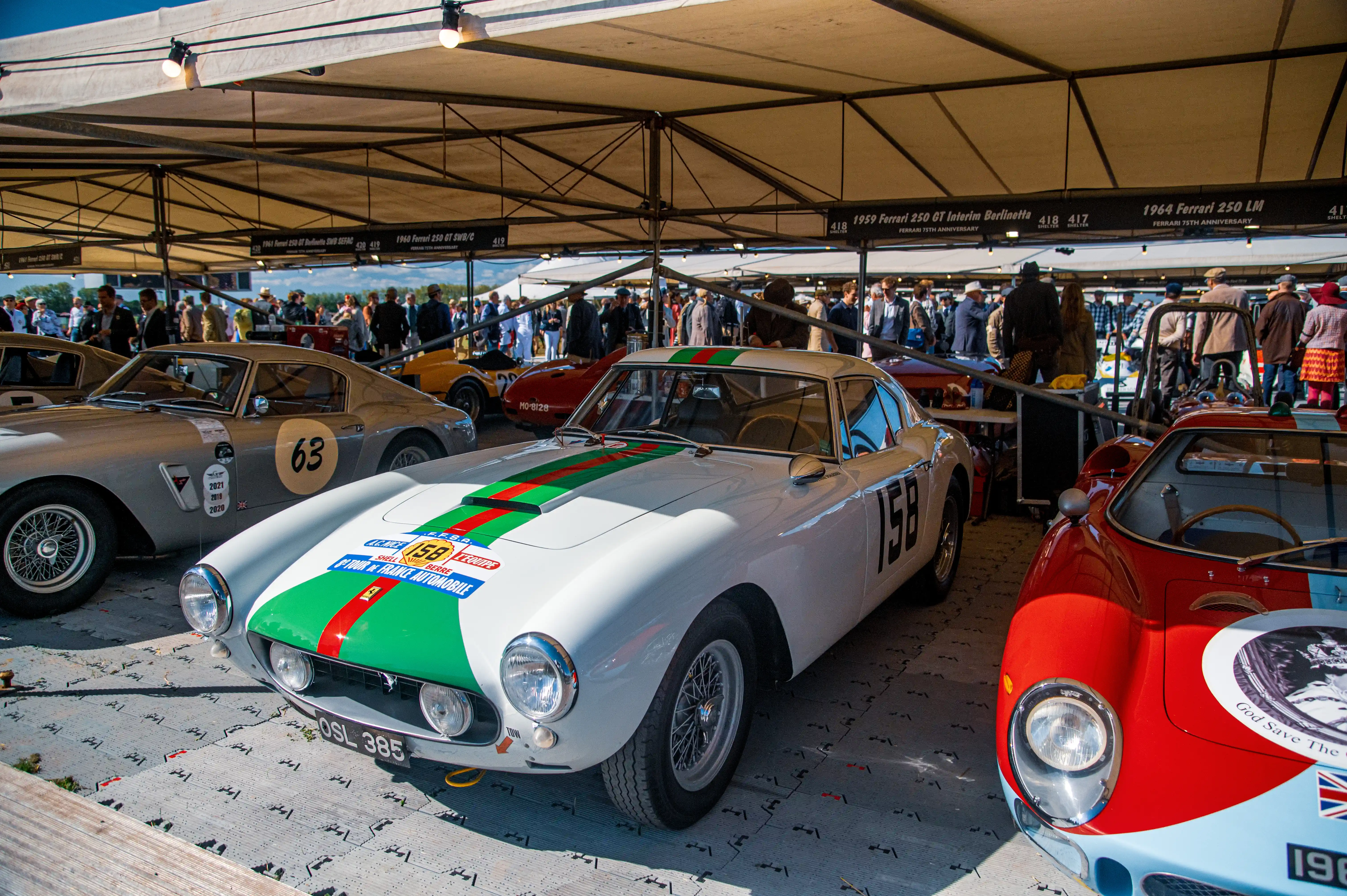 A white classic Ferrari 250 GT with distinctive green and red racing stripes is parked in the foreground. It has decals on its bonnet indicating participation in the 