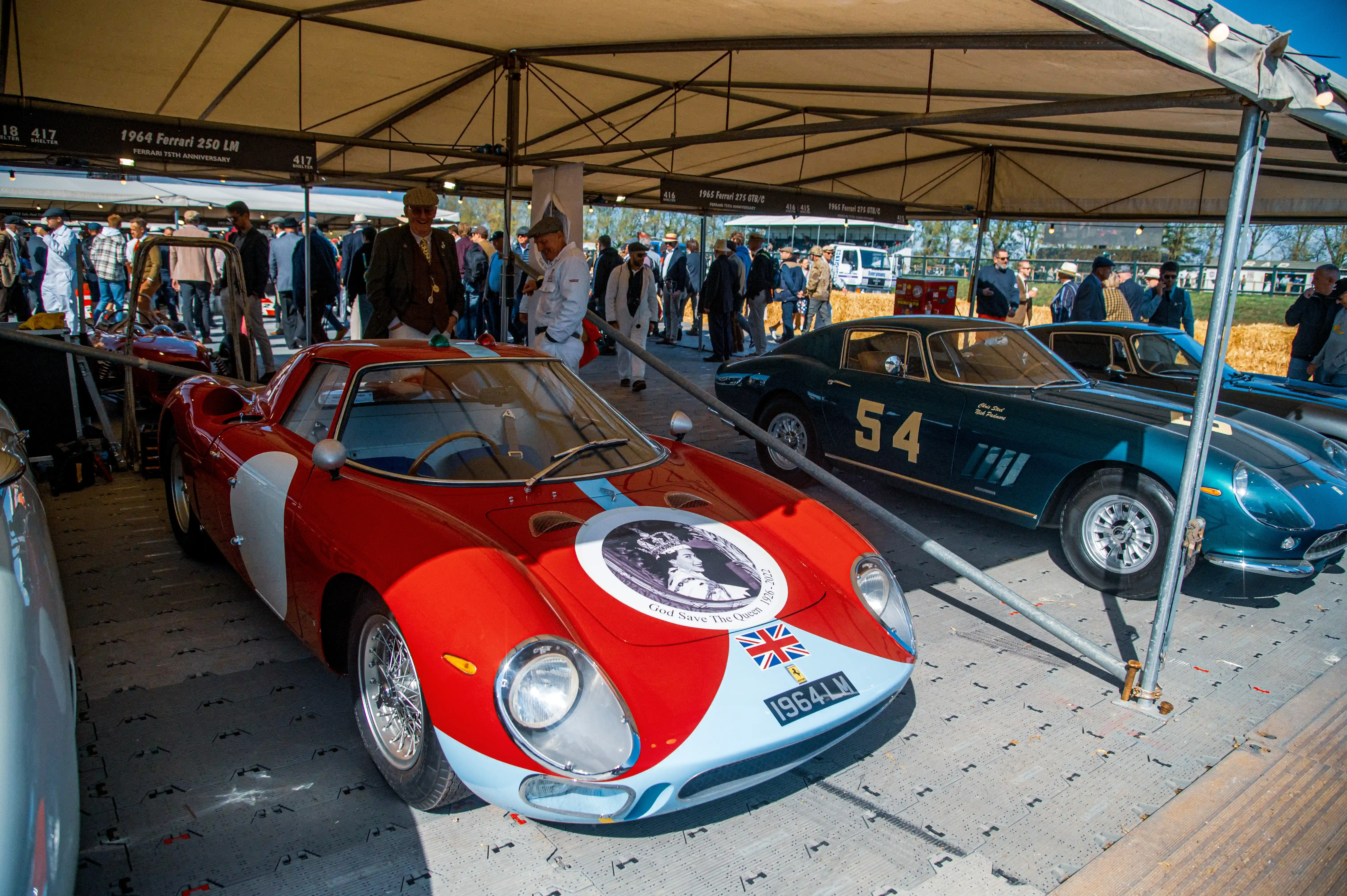 Close-up of a red 1964 Ferrari 250 LM with white accents and a depiction of Queen Elizabeth II on the bonnet, parked next to a dark green 1965 Ferrari 275 GTB/C with the number '54' on its side. People are visible in the background under a covered display area.