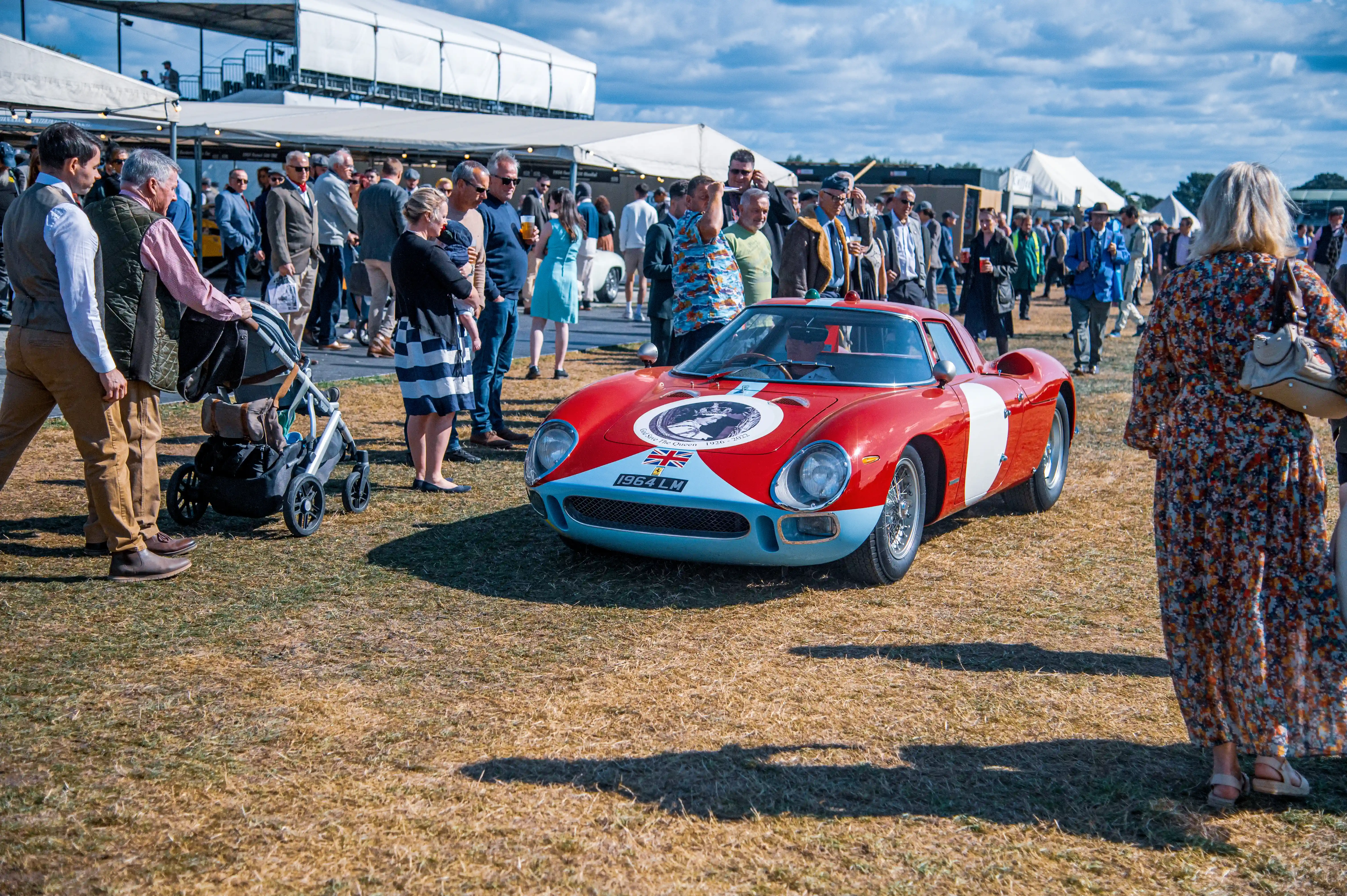 A classic red Ferrari sports car with blue accents and the number plate '1964 LM' is parked on a dry grass field. A diverse crowd of people, some holding drinks and babies, are gathered around the car. The car displays a tribute to Queen Elizabeth II and a Union Jack flag.