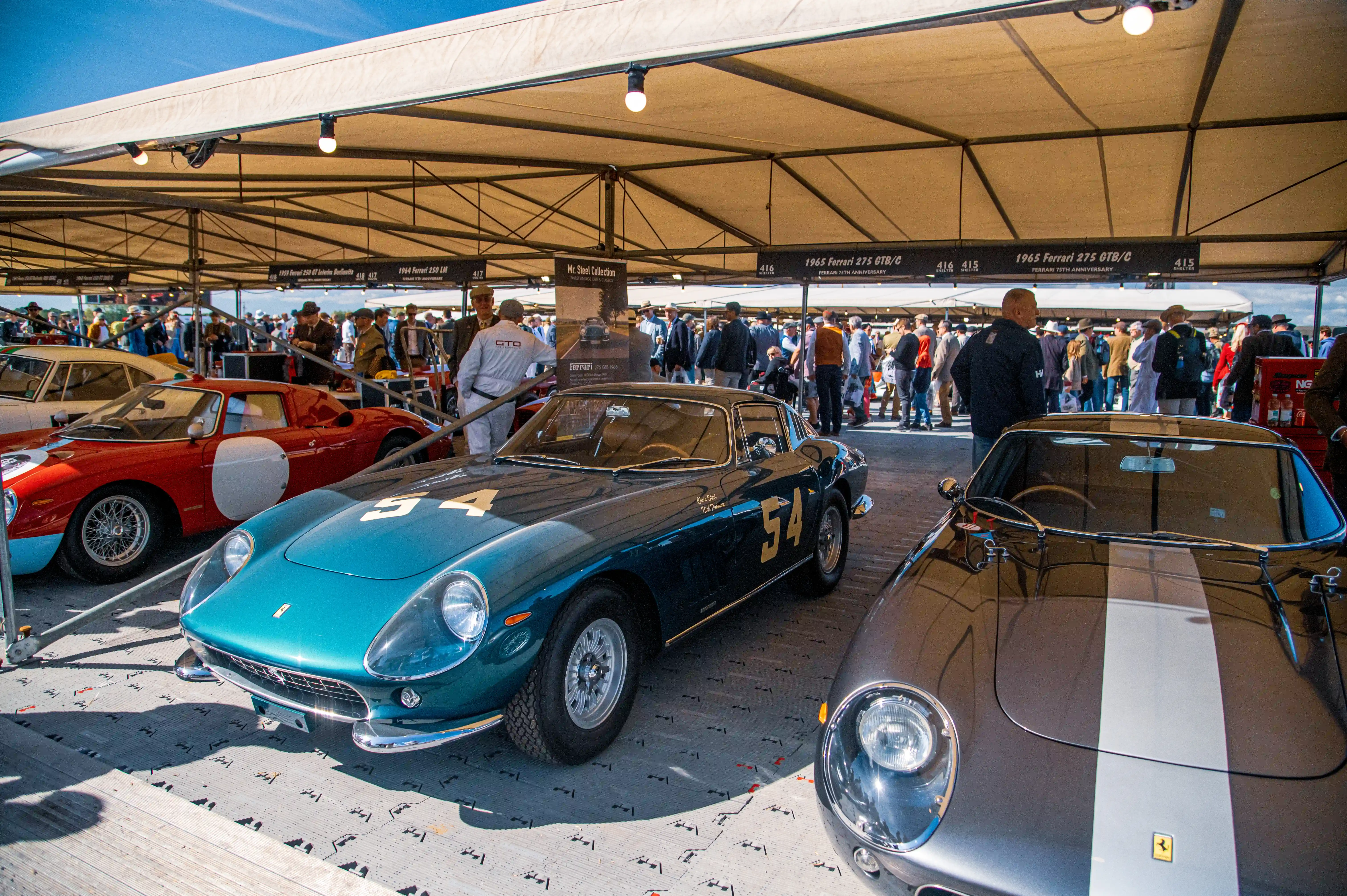 A vintage teal Ferrari 275 GTB/C, number 54, is parked next to a grey Ferrari with a white stripe under a tent at an outdoor event. Spectators are visible in the background.