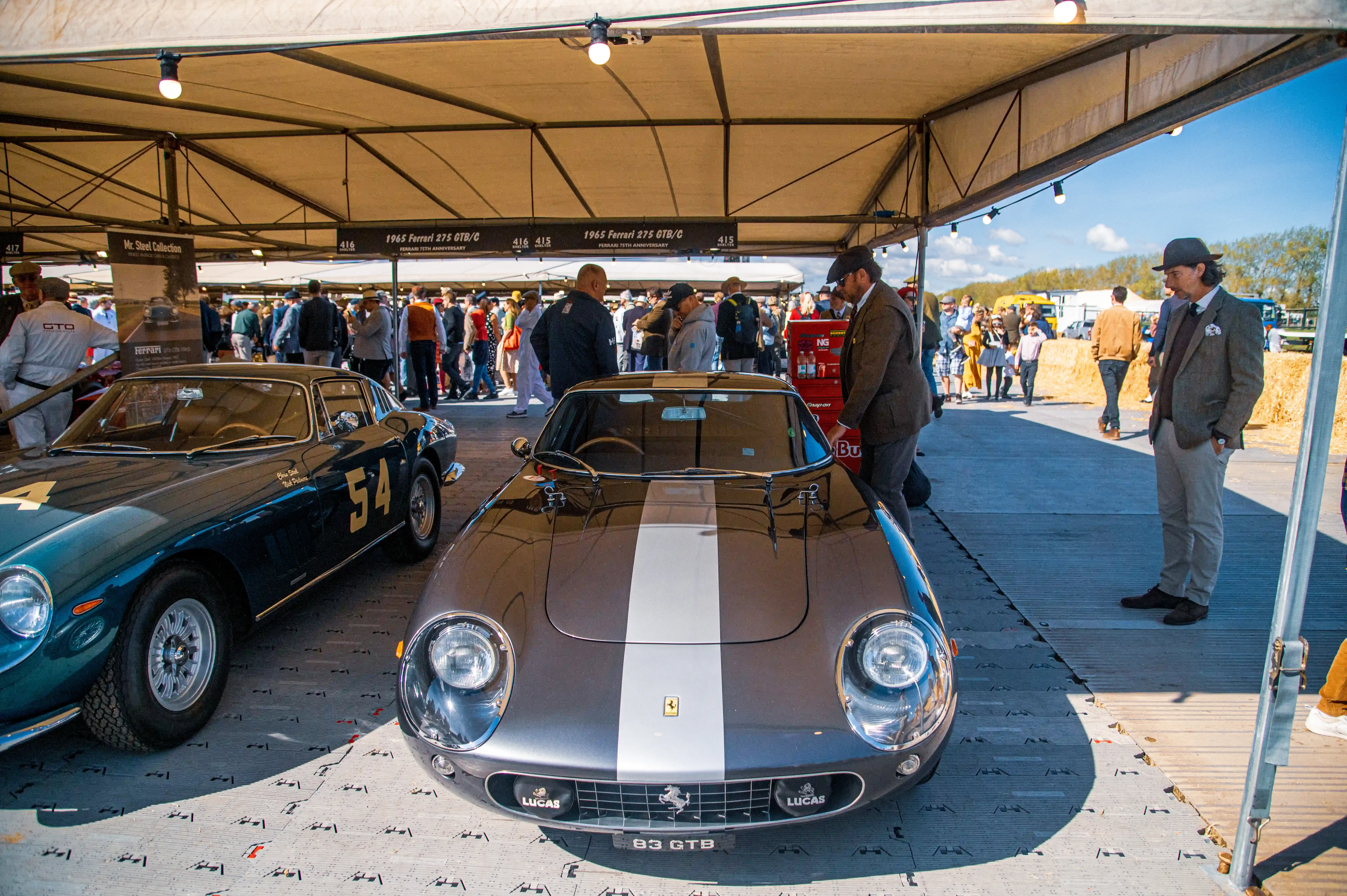 A grey 1965 Ferrari 275 GTB/C with a white stripe down the bonnet and two vintage Ferraris are displayed under a tent at an outdoor event. People are gathered around looking at the cars, and a man in a tweed jacket and hat is standing beside the grey Ferrari.