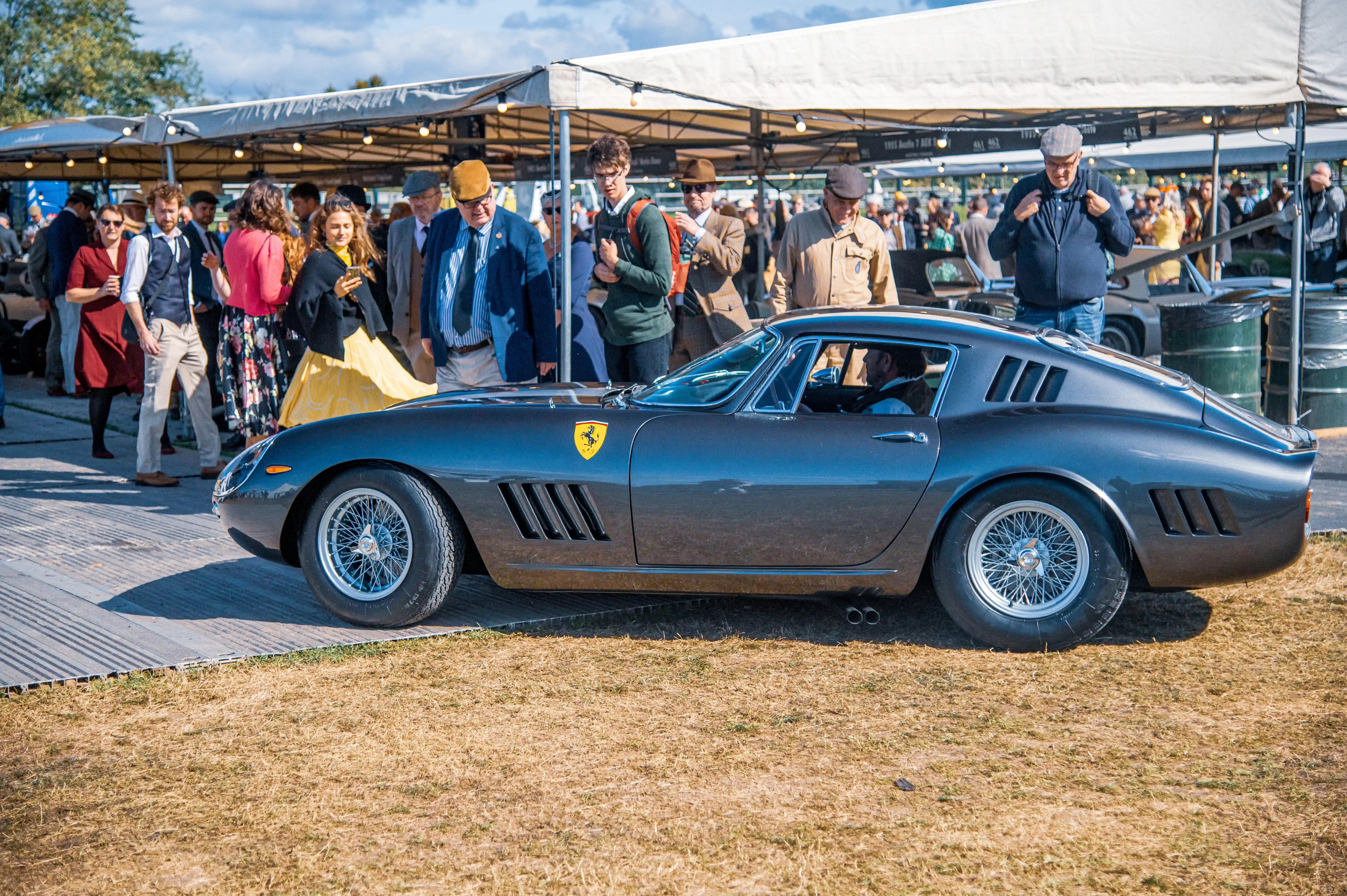 A classic dark grey Ferrari GT car with wire-spoke wheels is parked on grass. A crowd of people in smart casual and formal attire are gathered around it, looking at the vehicle under a white marquee.