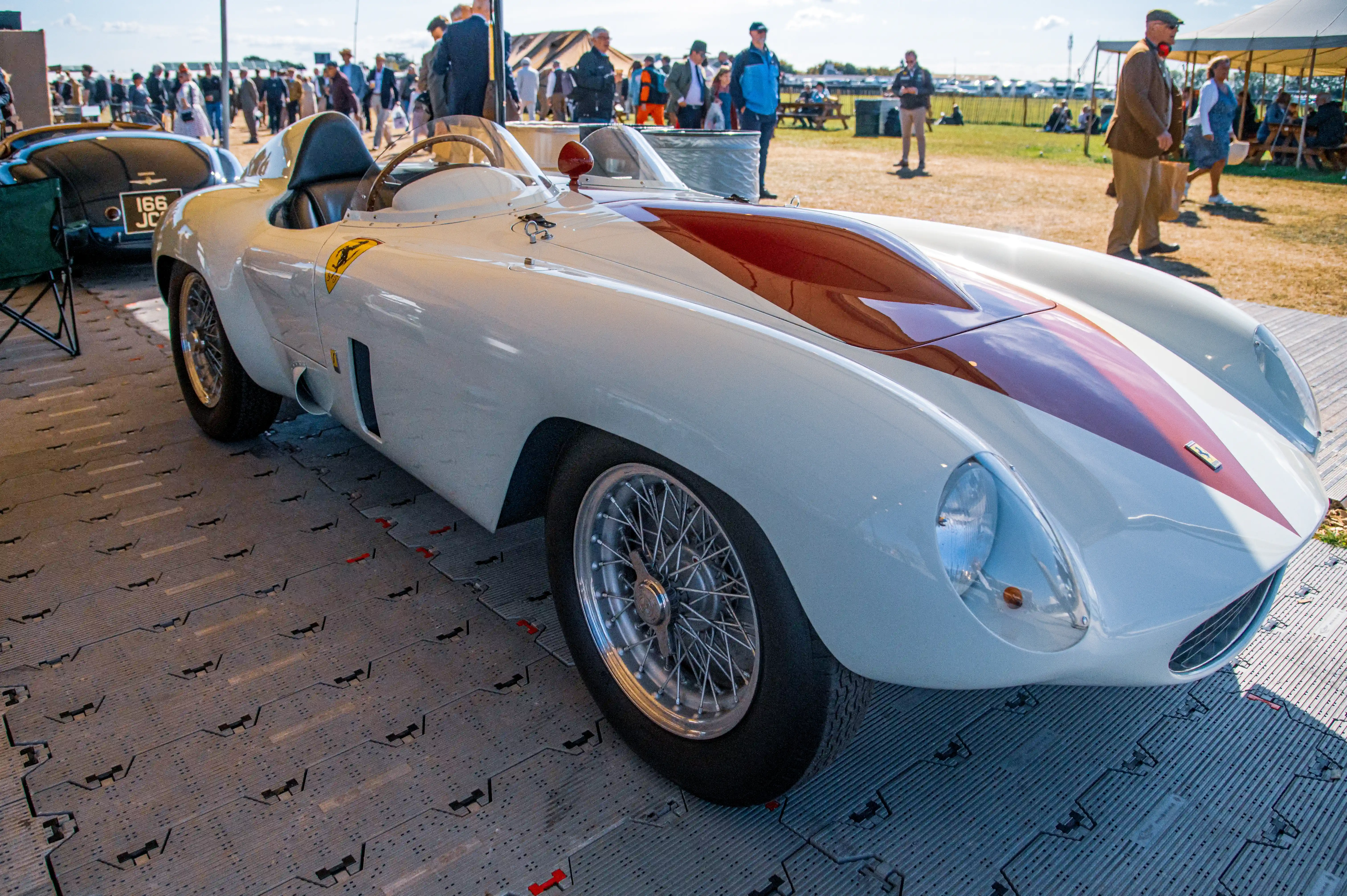 Close-up view of a classic white and maroon Ferrari sports car, featuring wire-spoke wheels and a low-profile windscreen. The car is parked on a grey interlocking floor panel system, with a blurred background of people and tents.