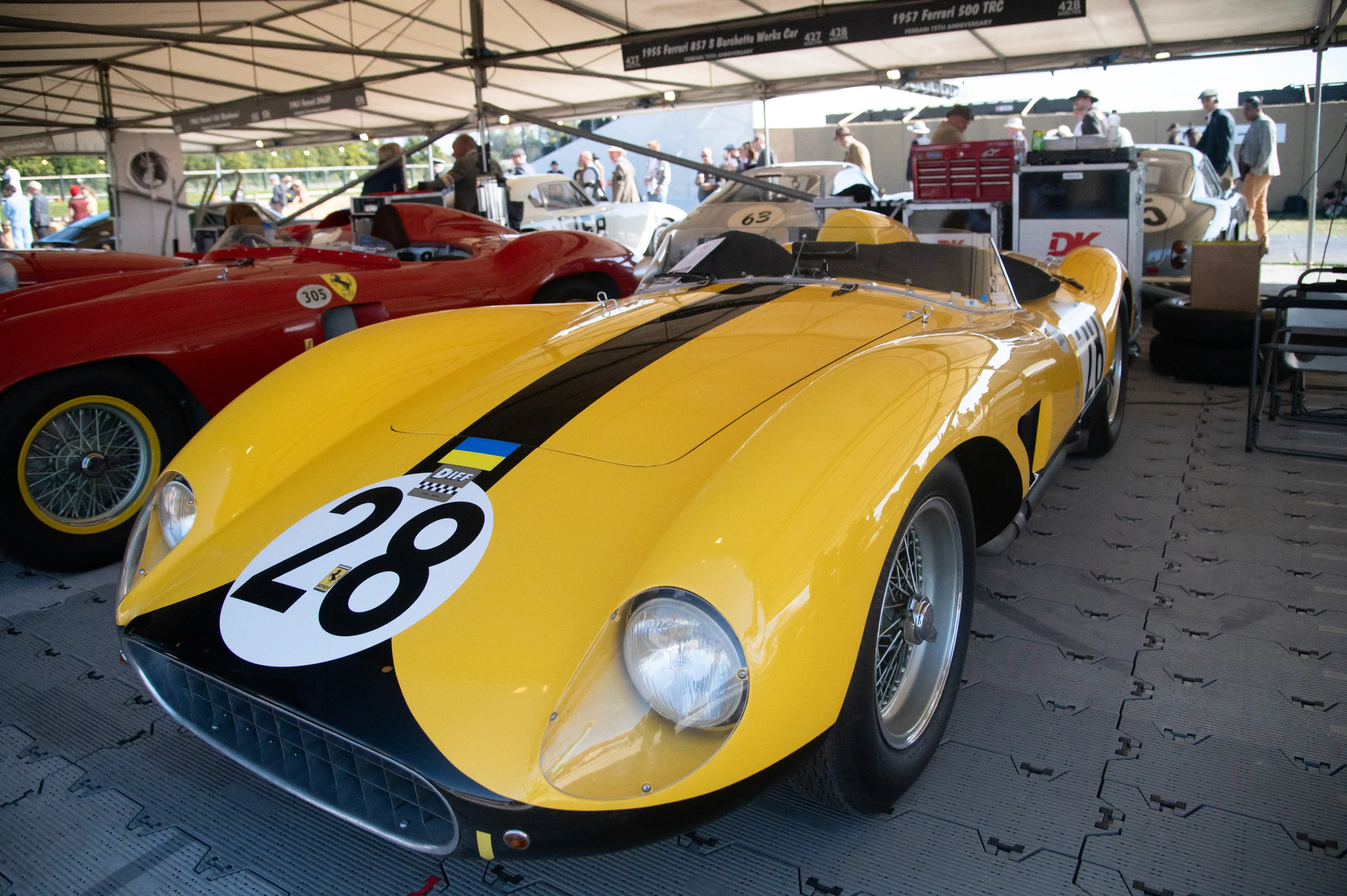 A close-up of a gleaming yellow vintage Ferrari race car with a black stripe and the number 28 in a white circle on its bonnet. A red vintage Ferrari is partially visible to the left.