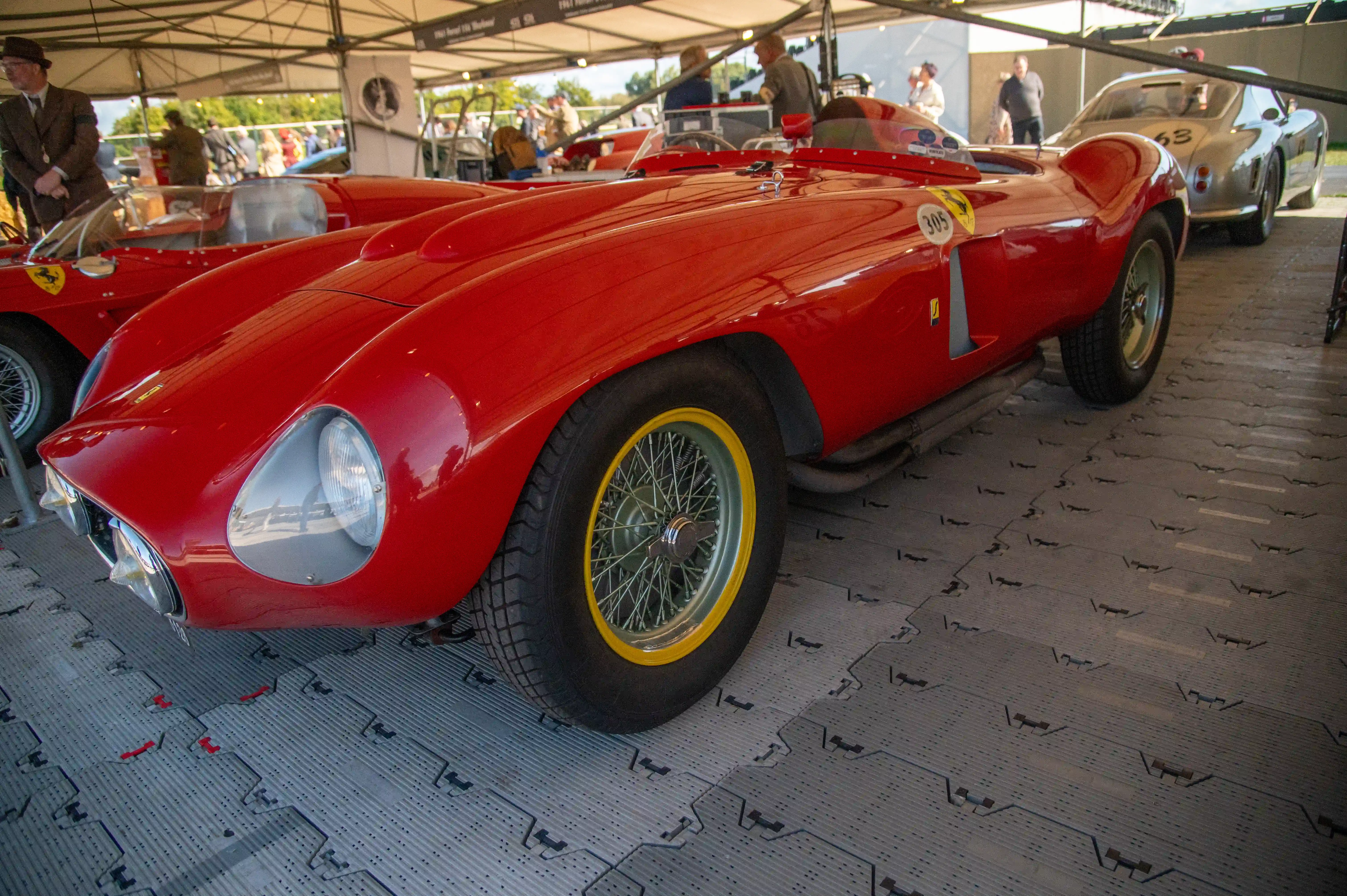  A classic dark grey Ferrari GT car with wire-spoke wheels is parked on grass. A crowd of people in smart casual and formal attire are gathered around it, looking at the vehicle under a white marquee.