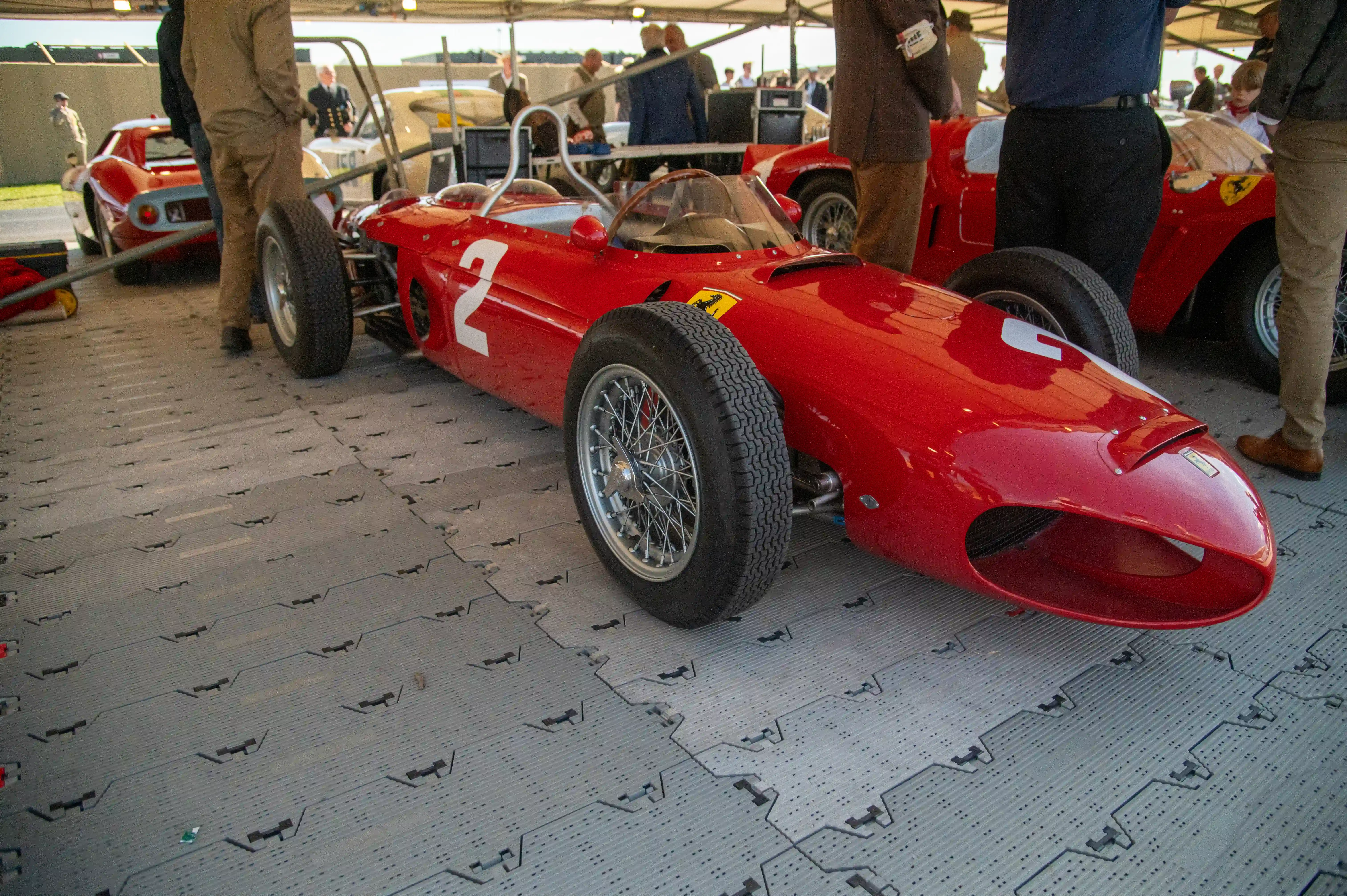A vintage red Ferrari Formula 1 car with the number 2 prominently displayed in white is parked on a grey modular floor. The car features a classic open-wheel design and wire-spoke wheels. People and other classic cars are visible in the soft-focus background