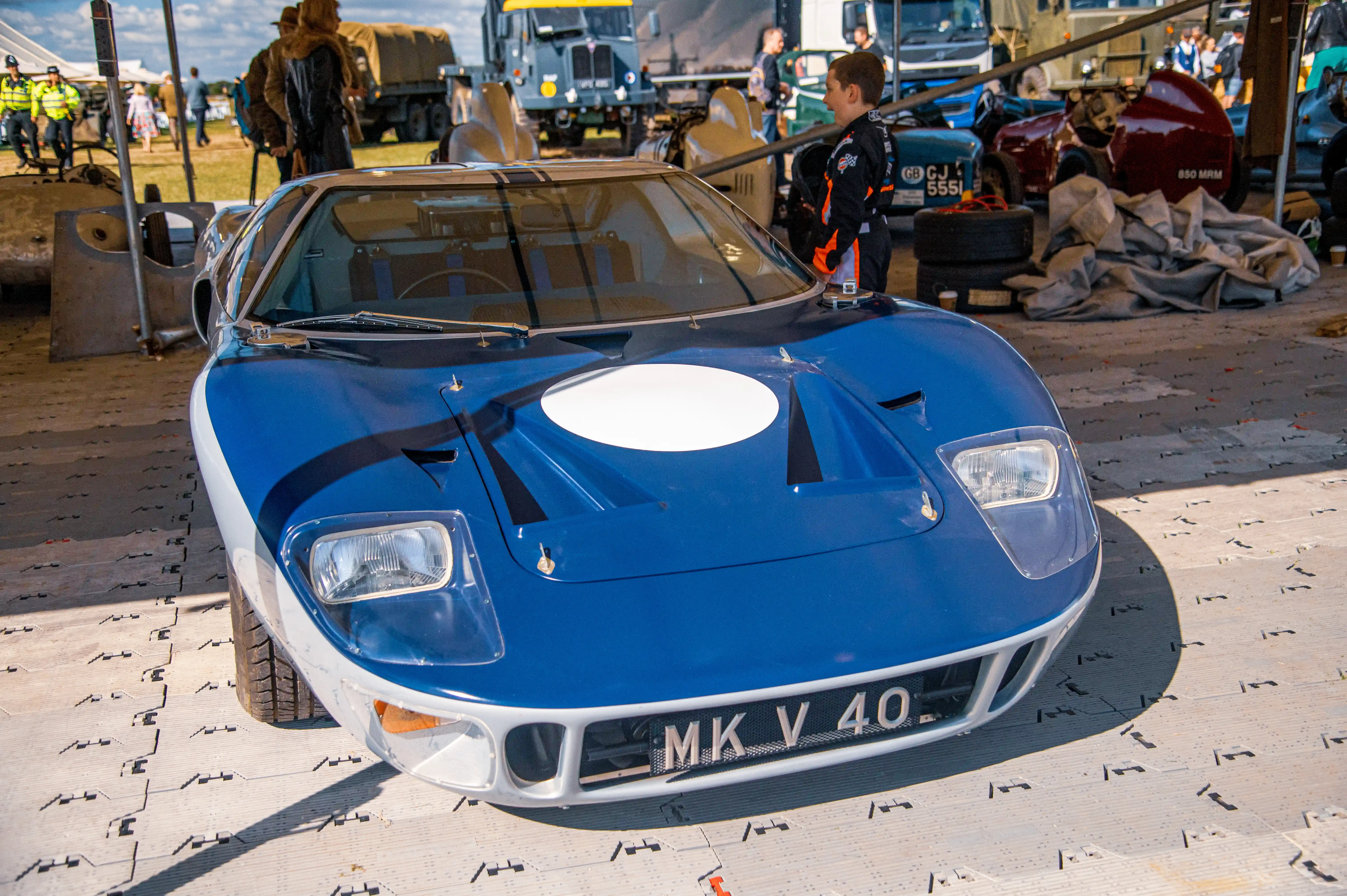 Front view of a blue and white Ford GT40 race car with a white circular decal on the bonnet, parked on a textured surface. A young boy in a racing suit stands to the right of the car.