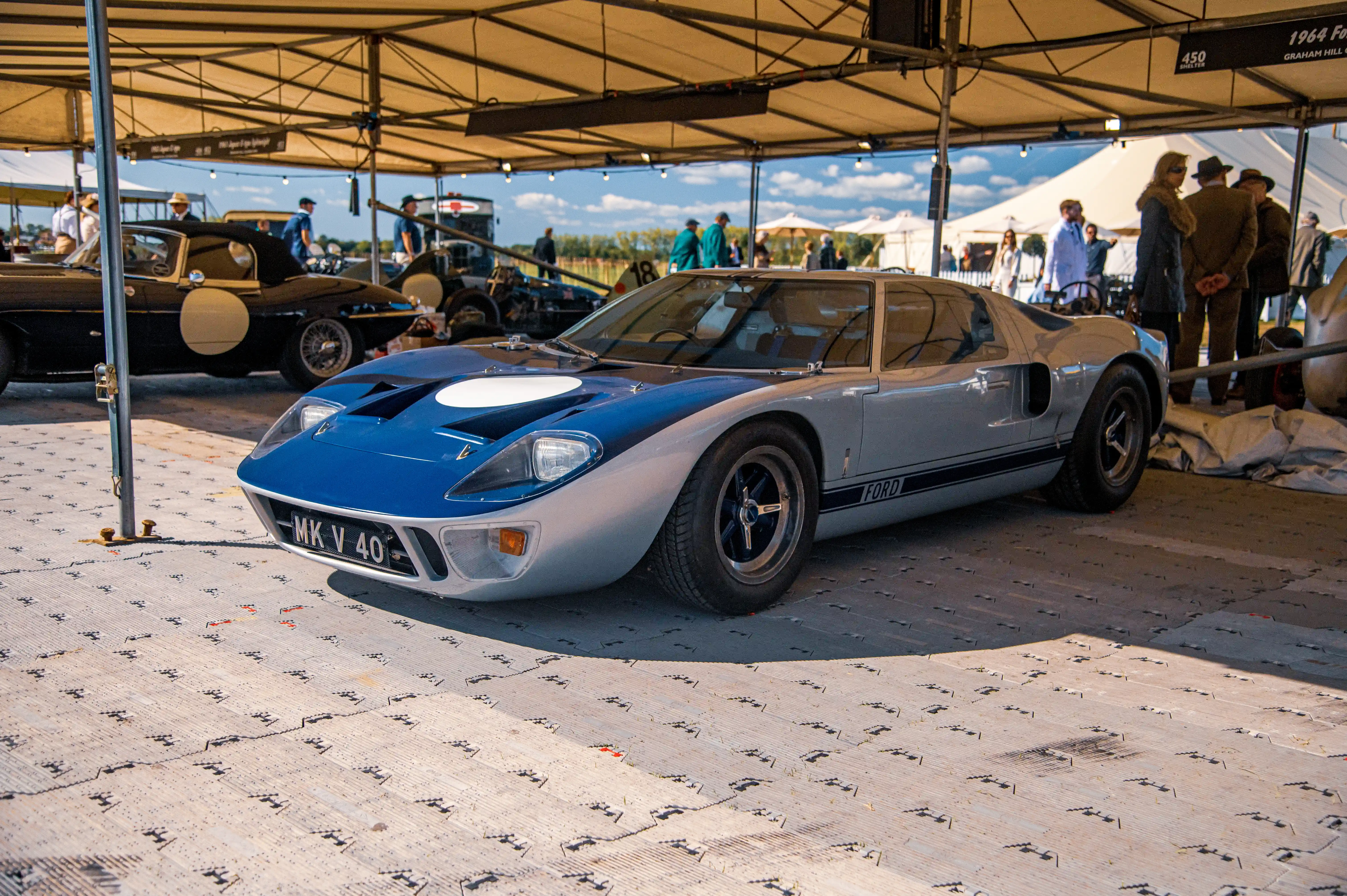 Front view of a blue and white Ford GT40 race car with a white circular decal on the bonnet, parked on a textured surface. A young boy in a racing suit stands to the right of the car.