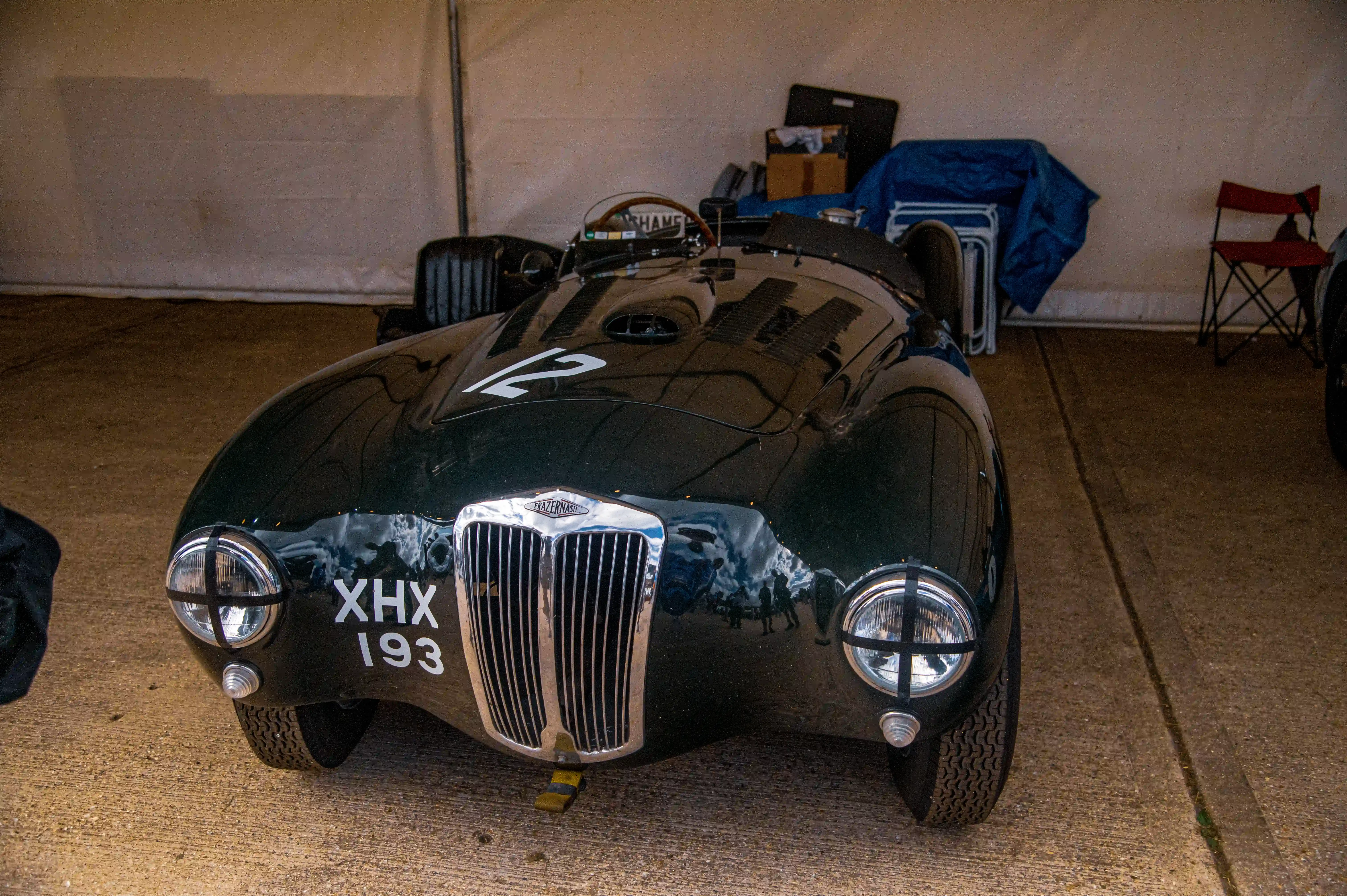 A front-three-quarter view of a dark green classic racing car with registration XHX 193 and racing number 12, parked indoors on a concrete floor.