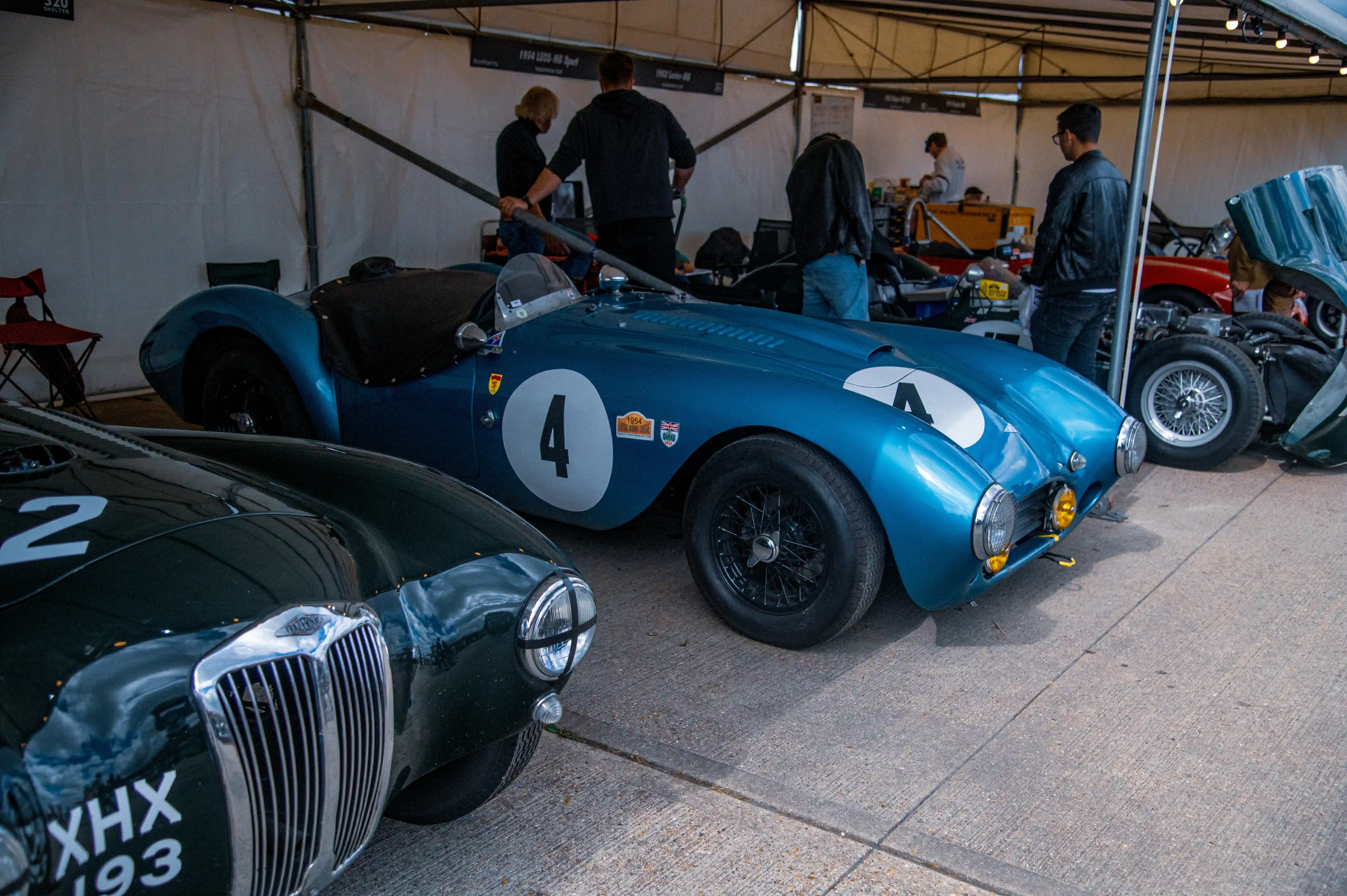 Two classic British sports cars, a blue one with a large number '4' on its side and a dark green one with a grille and number '2', are parked side-by-side indoors at an exhibition.