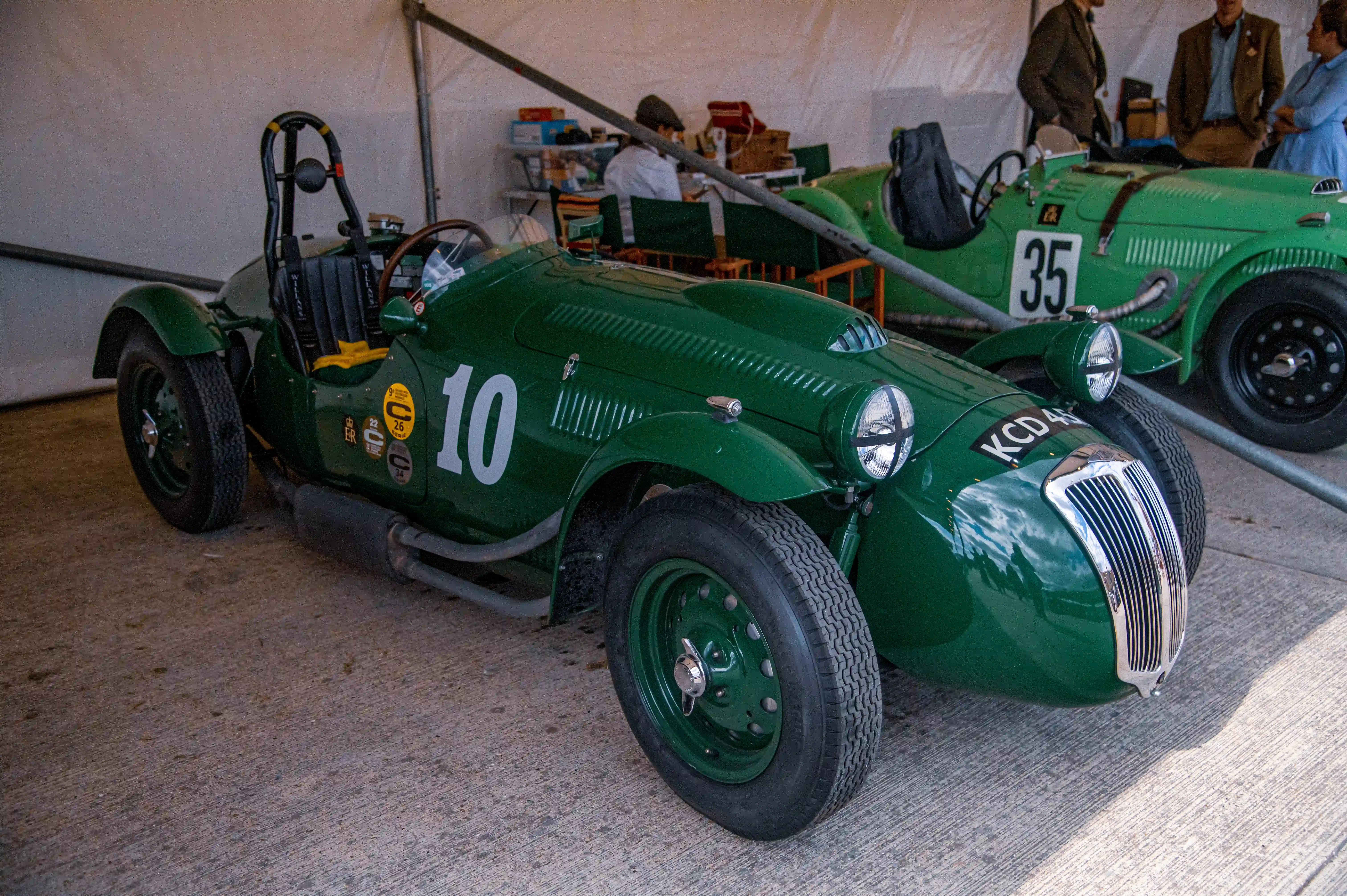 A close-up of a vintage green single-seater racing car with the number 10 on its side, parked next to another green racing car with the number 35. The cars are under a tent, and people in the background are dressed in vintage clothing.