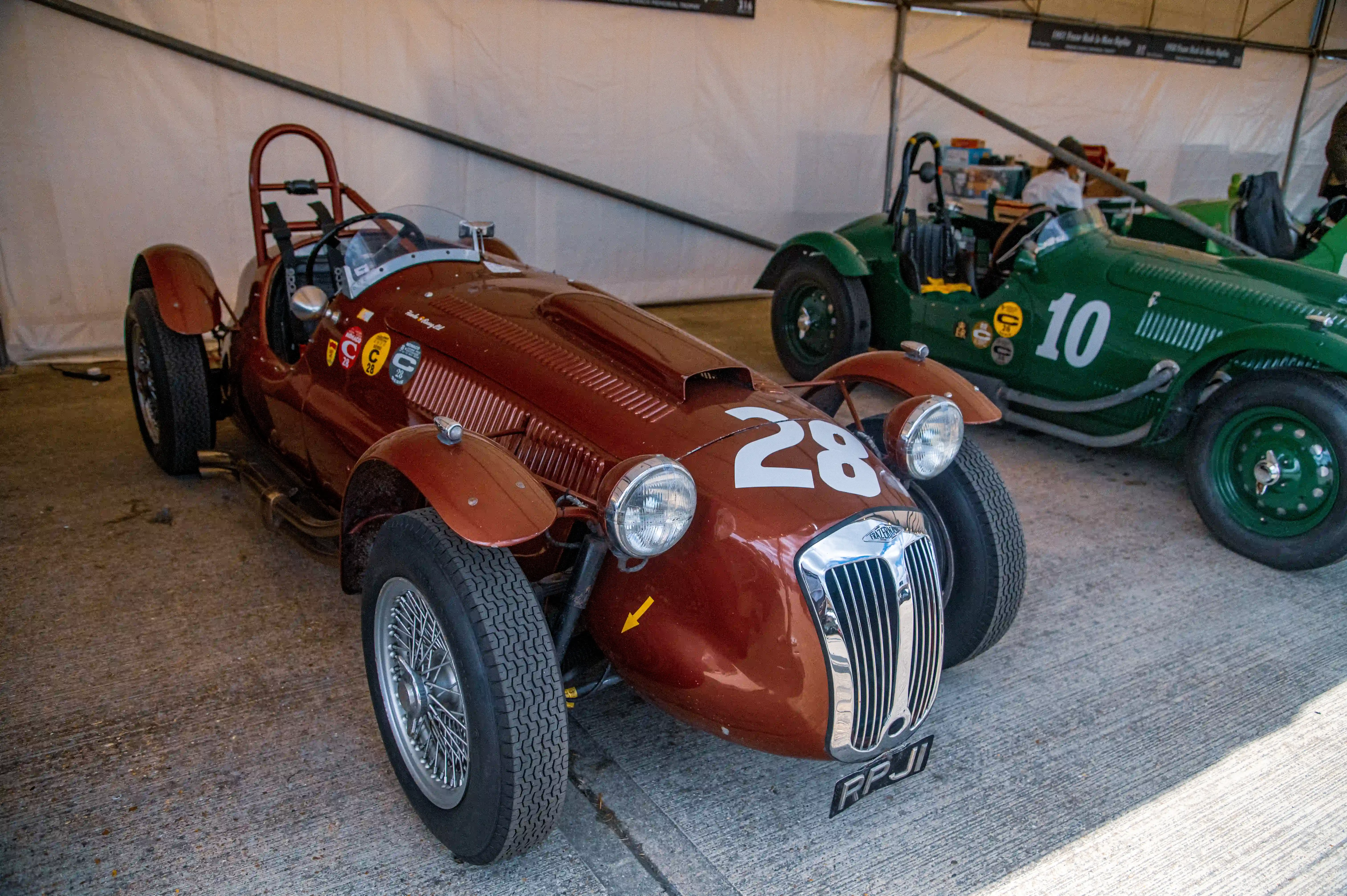 Two vintage open-top racing cars, one brown with number 28 and the other green with number 10, are parked next to each other.