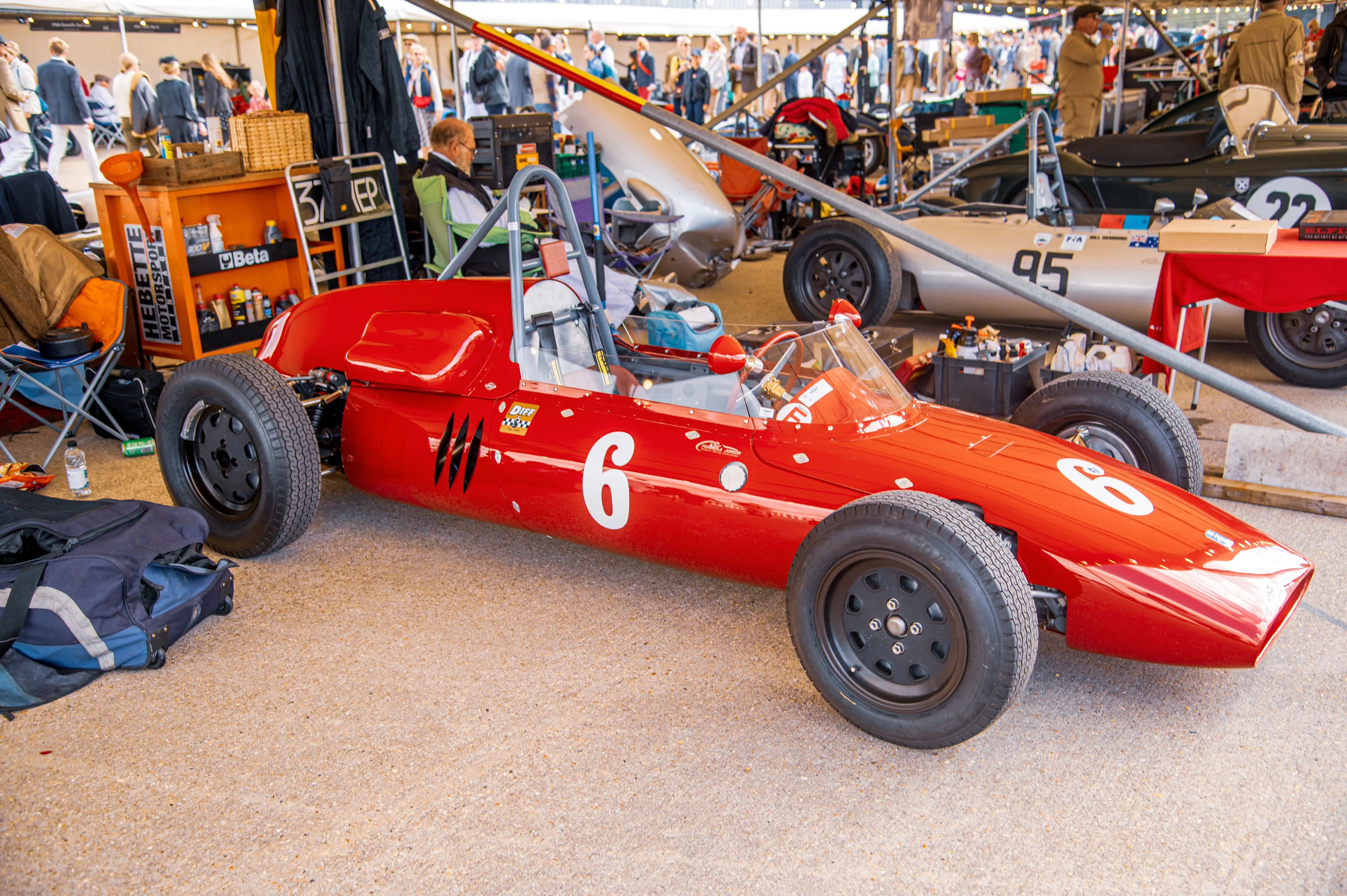A bright red vintage Formula 1 style racing car with the number 6 in white on its side is parked on a light-coloured, textured ground. The car has black wheels and an open cockpit. In the blurred background, there are other cars, people, and stalls.