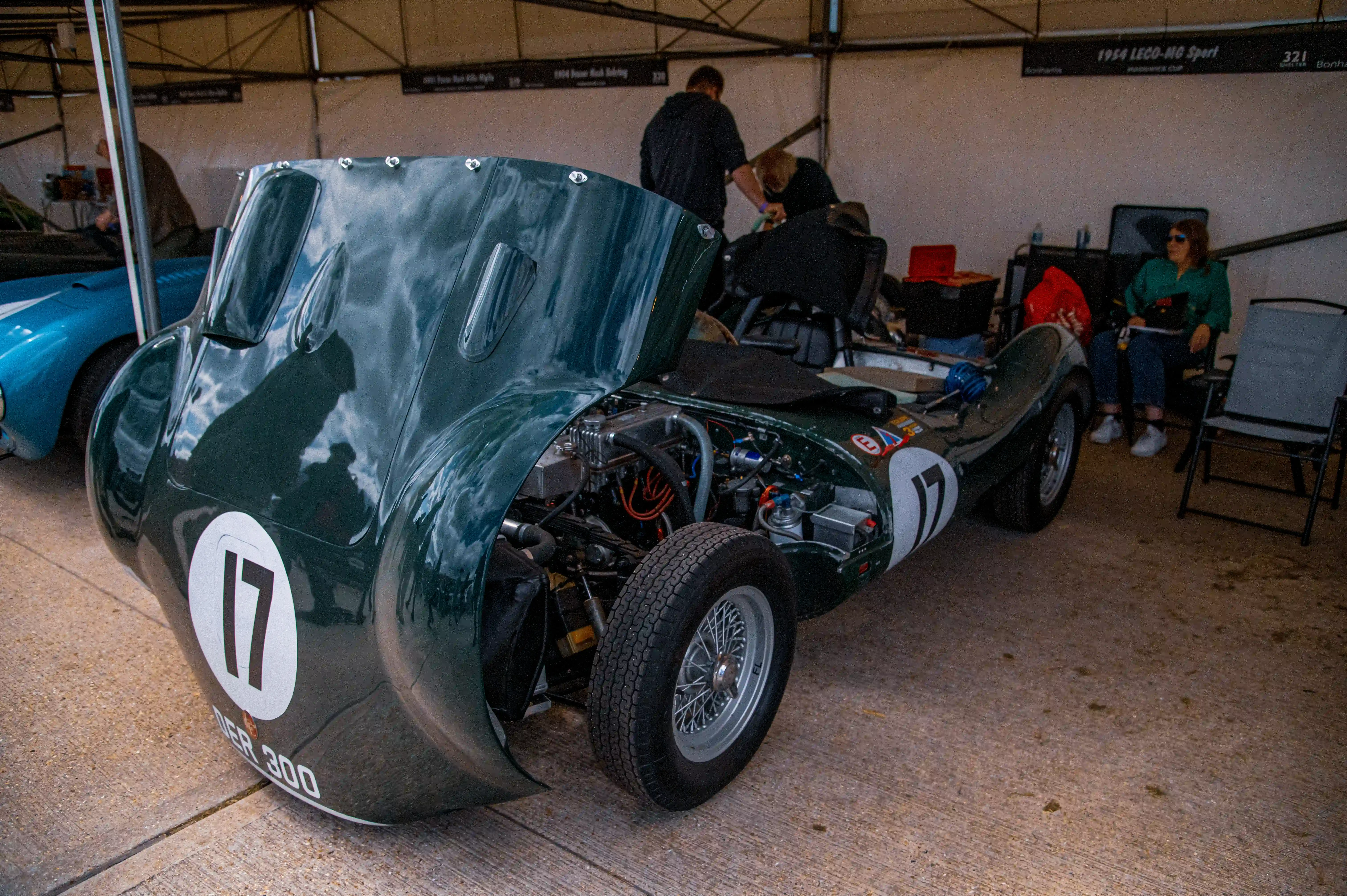 A close-up of a dark green vintage racing car with its bonnet lifted, showing the engine. The car has the number 17 in a white circle on its side and 