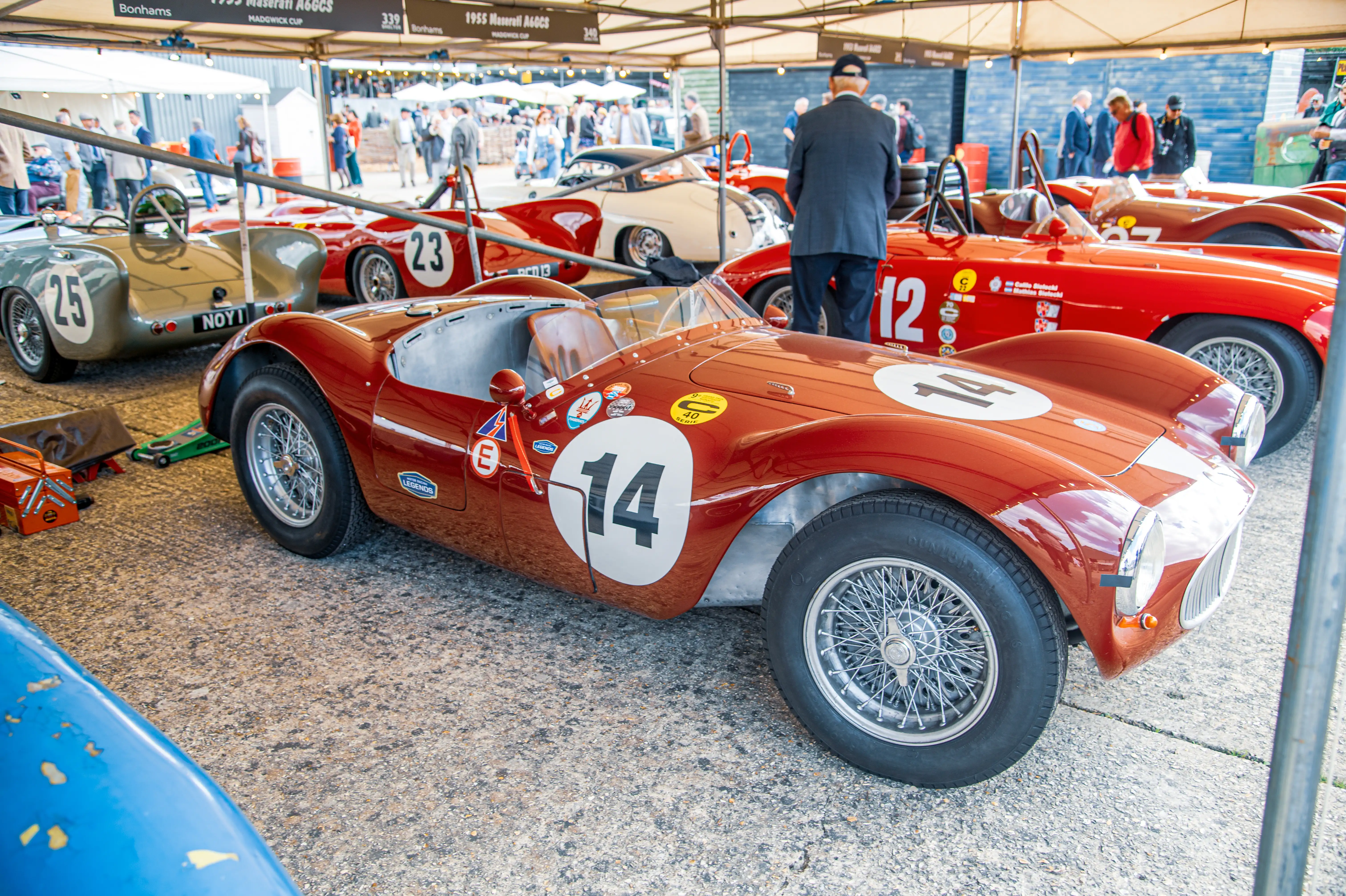 Three classic racing cars parked side-by-side under a tent. The car in the foreground is red with the number '14' on its side. To its left is a light grey car with the number '25'. Behind these are red cars with numbers '23' and '12'. People are visible in the background.