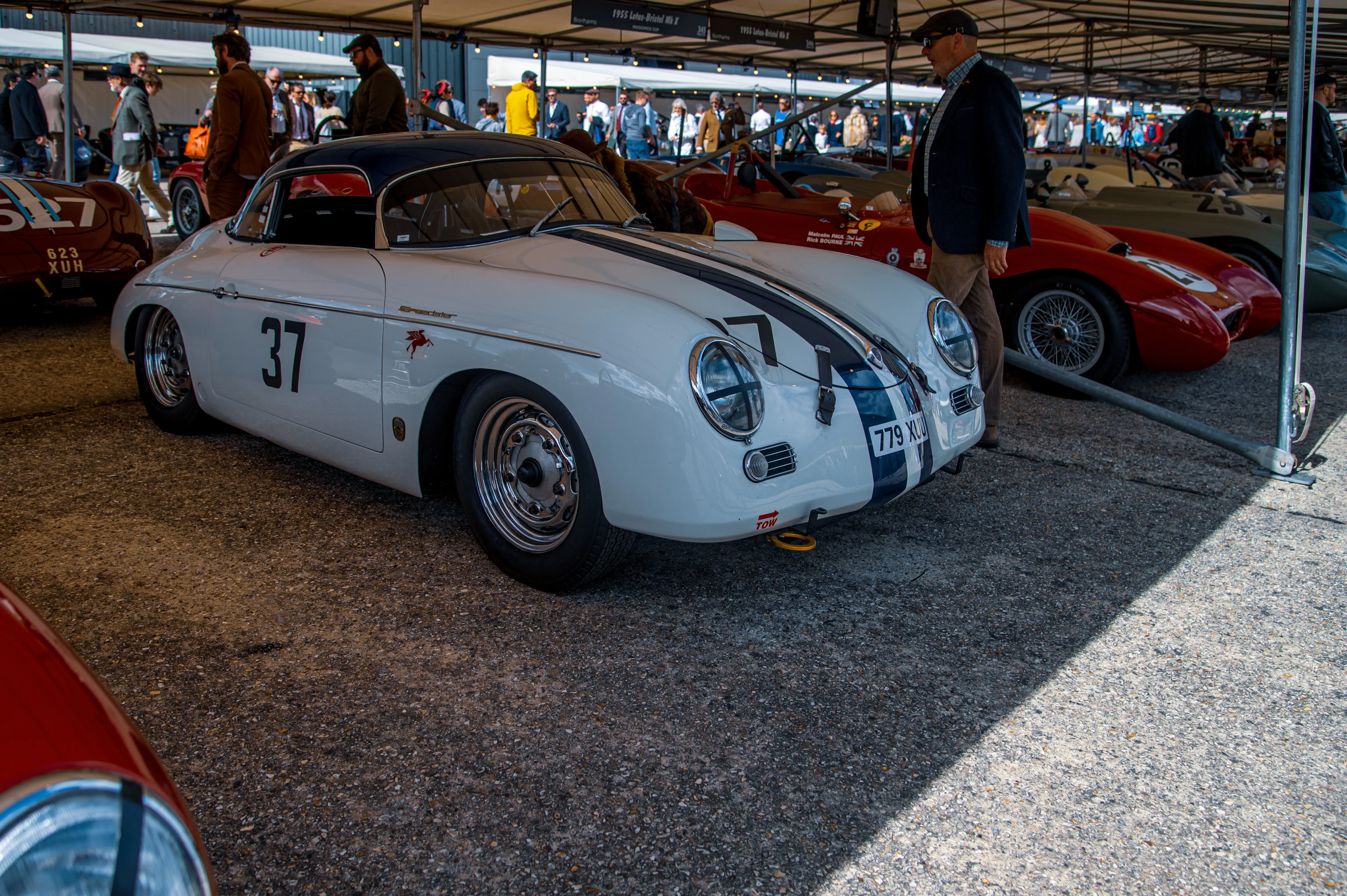 A close-up, low-angle shot of a vintage white Porsche Speedster with dark blue racing stripes and the number '37' on its side. The car is parked on a gravel surface under a canopy, with other classic cars and people in the background.