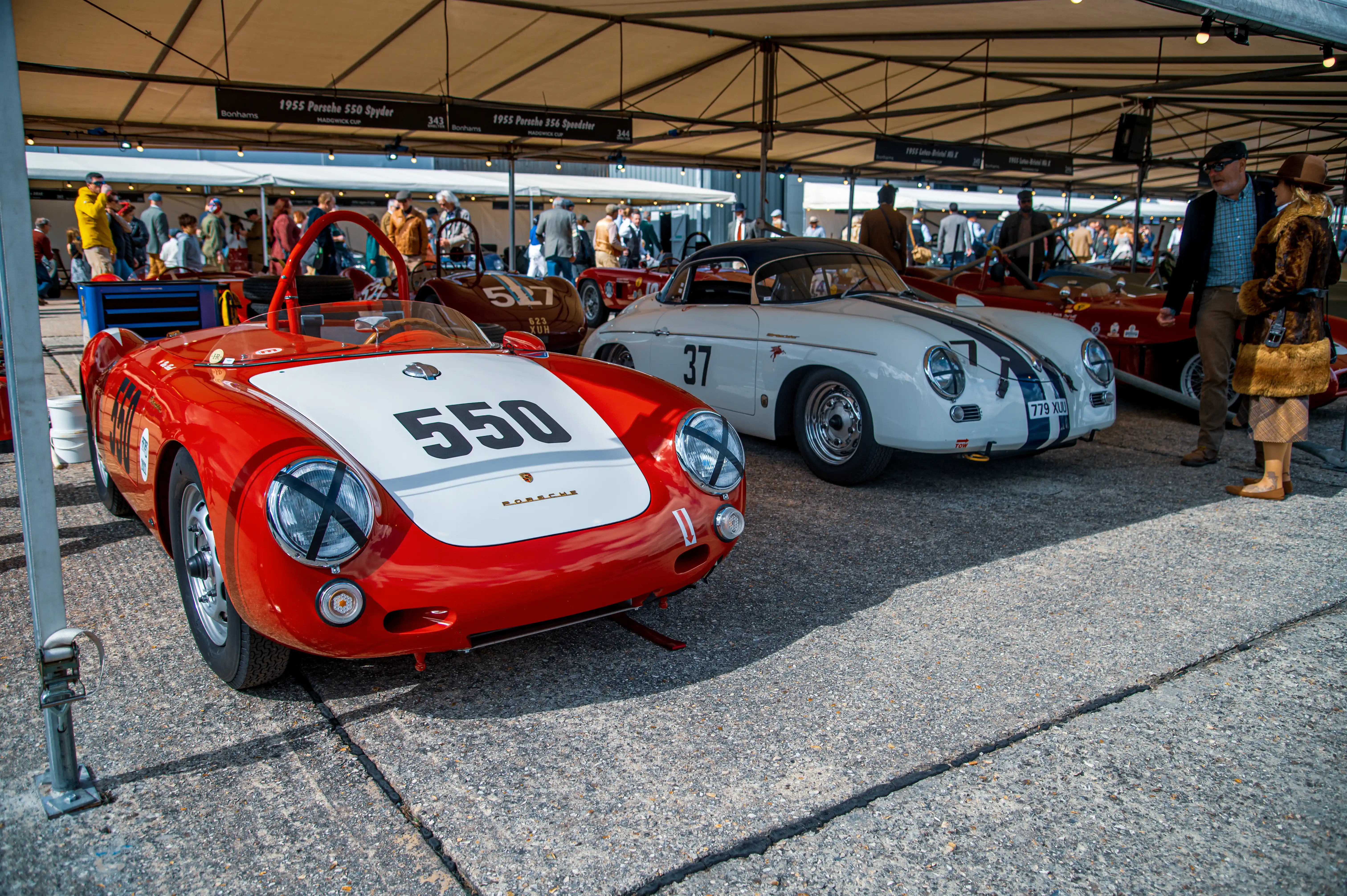 Close-up of a red 1955 Porsche 550 Spyder with white accents and the number '550' on its bonnet, parked next to a white 1955 Porsche 356 Speedster with blue stripes and the number '37' on its door. Several people are visible in the background, looking at vintage cars displayed under a tent at an outdoor event.