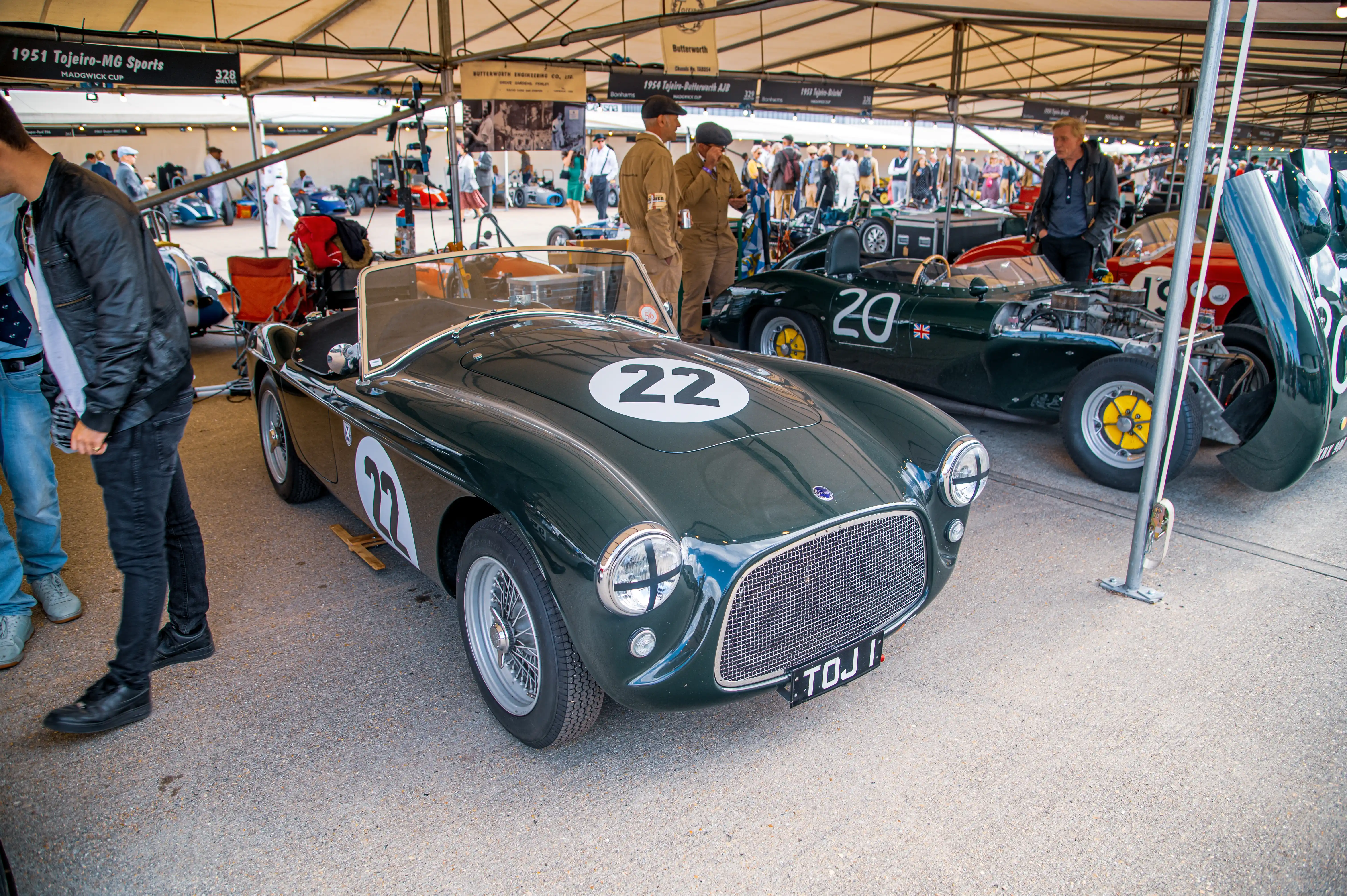 A dark green classic sports car with the number 22 on its bonnet is parked in the foreground, with another dark green racing car numbered 20 and a red sports car visible behind it under a tent. Men in vintage racing outfits are present.