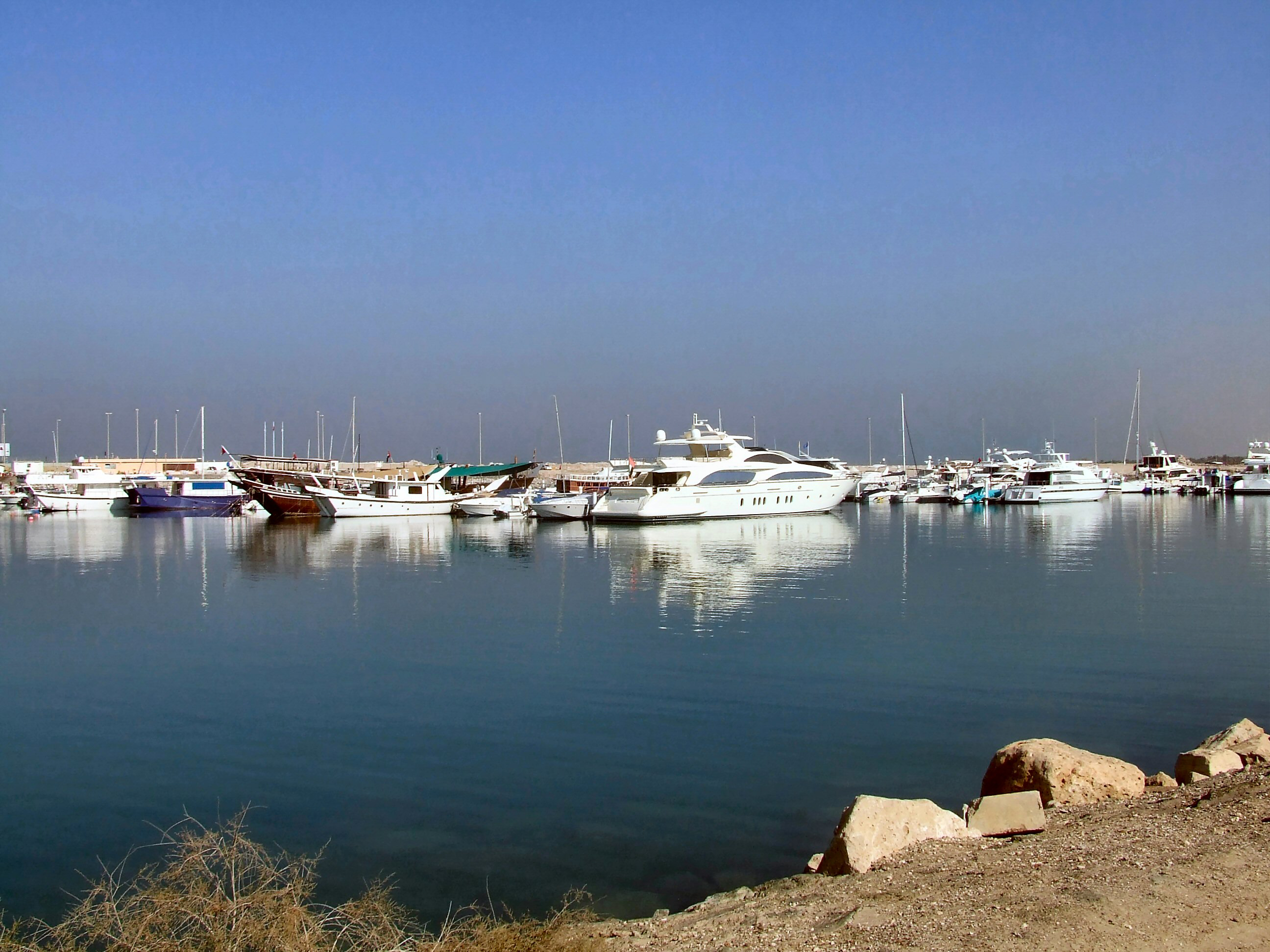 The image depicts a serene marina with various boats and yachts docked at the harbor. The water is calm, reflecting the boats and the clear blue sky above. The scene is tranquil, with a few large rocks in the foreground and a hazy background that suggests a distant shoreline or landmass.
