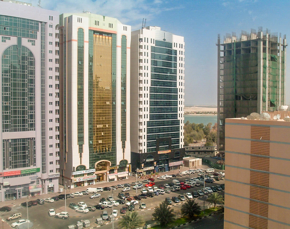 The image depicts a cityscape with several high-rise buildings, a busy street, and a parking lot filled with numerous cars. The buildings are modern and primarily composed of glass and concrete, reflecting a contemporary urban environment. The street below is lined with various commercial establishments, and the scene is set against a backdrop of a body of water and distant landscape.