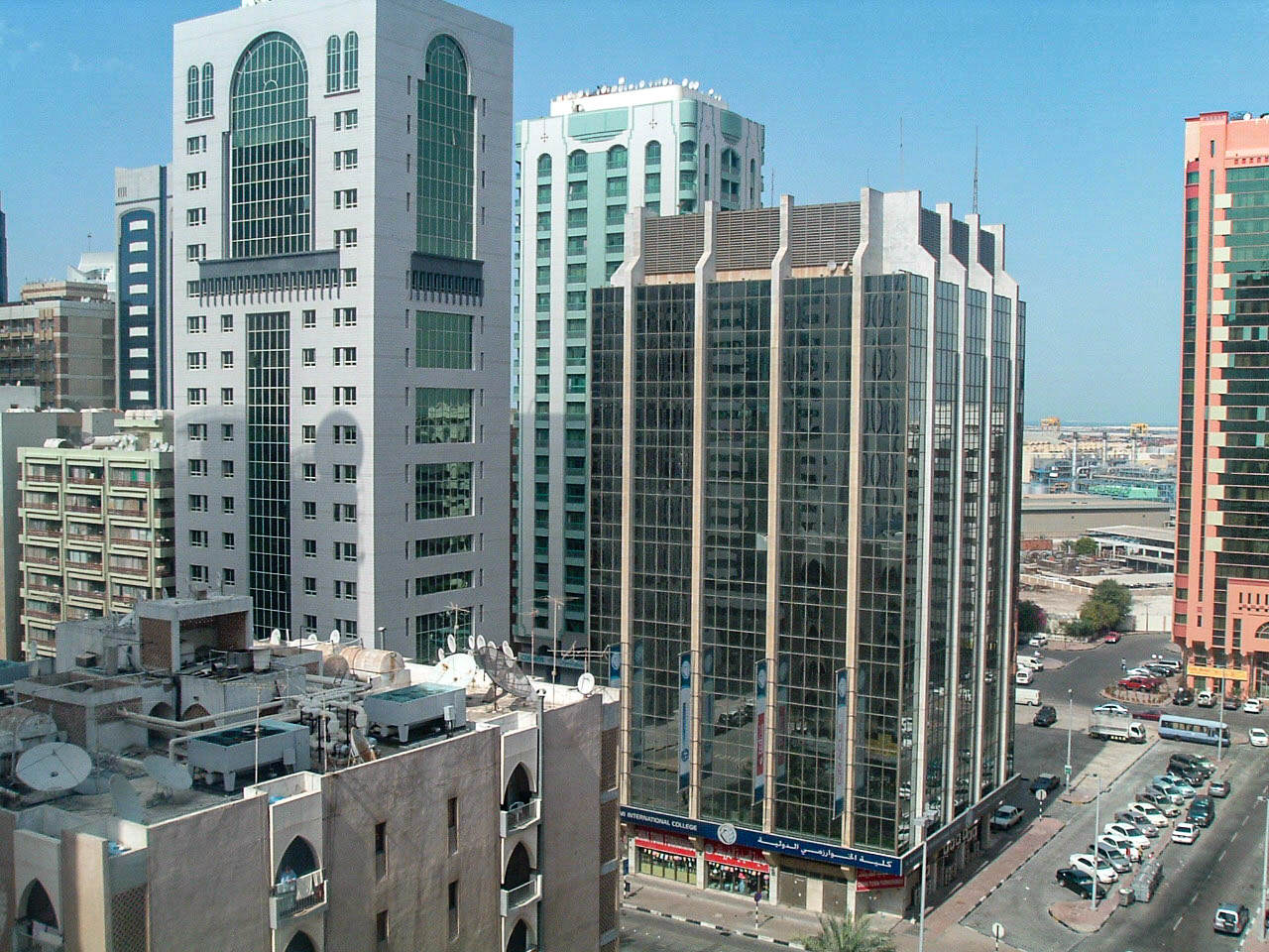 The image depicts a cityscape featuring several modern high-rise buildings with a mix of architectural styles. The buildings are primarily composed of glass and concrete, showcasing a blend of contemporary and traditional design elements. The scene includes a busy street with parked cars and some construction activity, indicating an urban environment. The sky is clear, suggesting a sunny day. The overall atmosphere is that of a bustling, developed city.